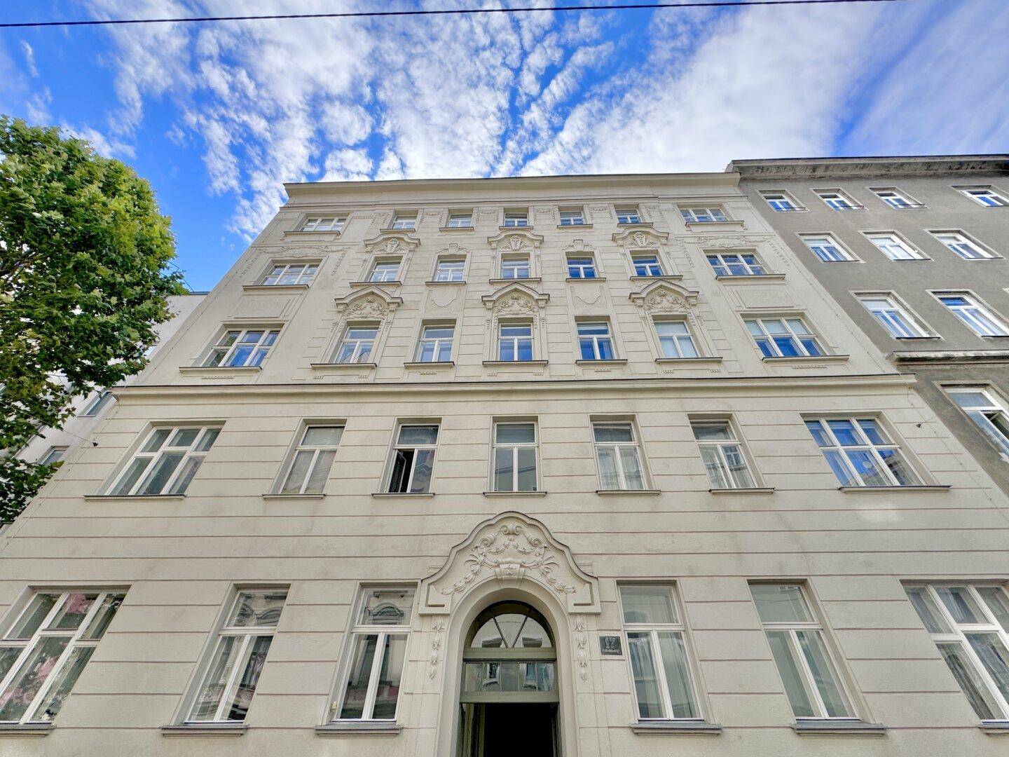 Historische Fassade eines Altbaus mit eleganten Verzierungen und großen Fenstern unter blauem Himmel.