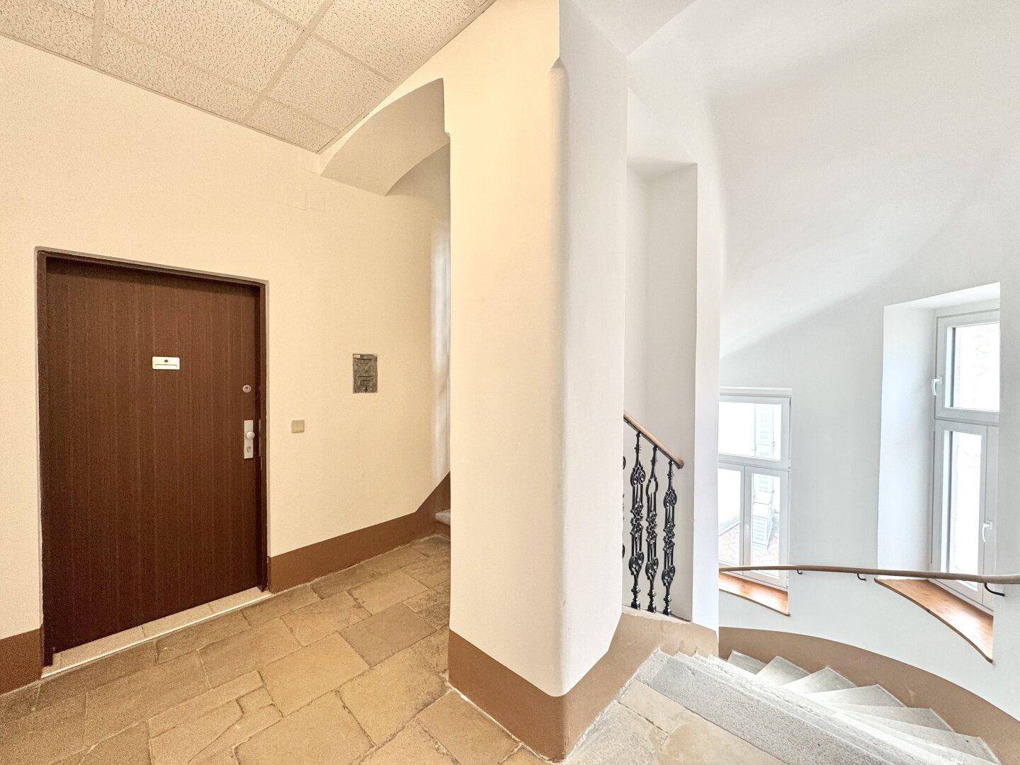 Heller Eingangsbereich mit Steinfliesenboden, Treppenaufgang und großen Fenstern im Treppenhaus.
