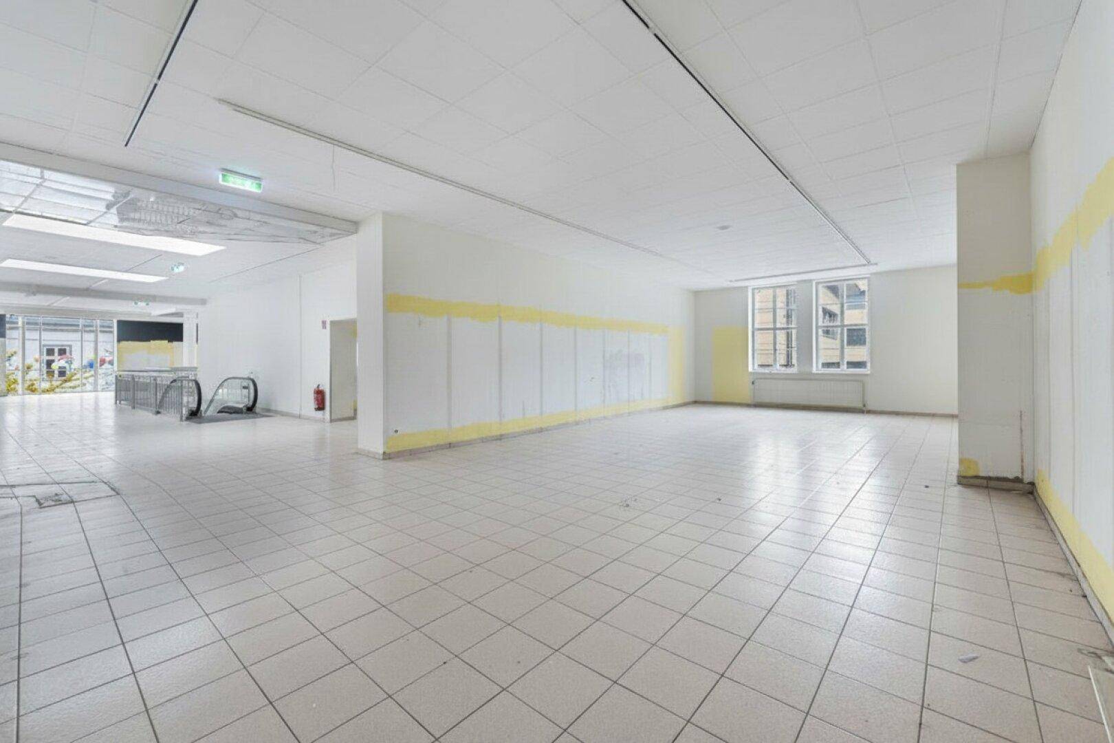 Langer, leerstehender Verkaufsraum mit Fliesenboden und gelben Farbakzenten an den Wänden.