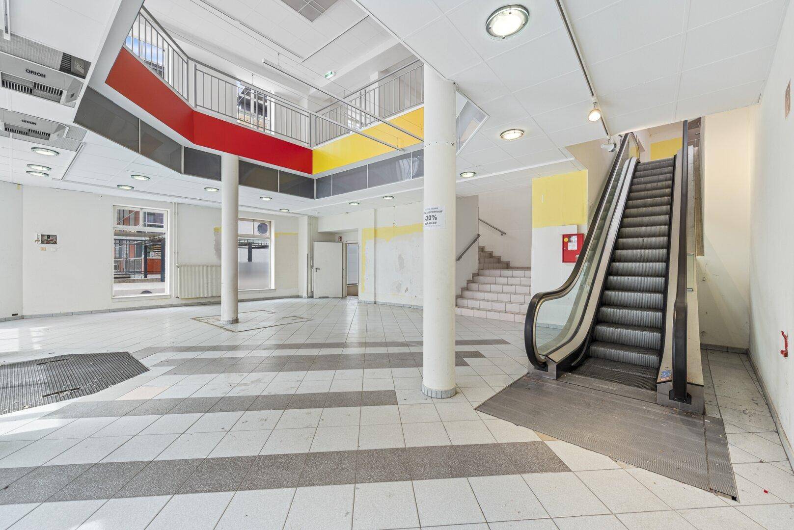 Heller Eingangsbereich mit Rolltreppe, Säulen und einem offenen Blick auf die oberen Etagen.
