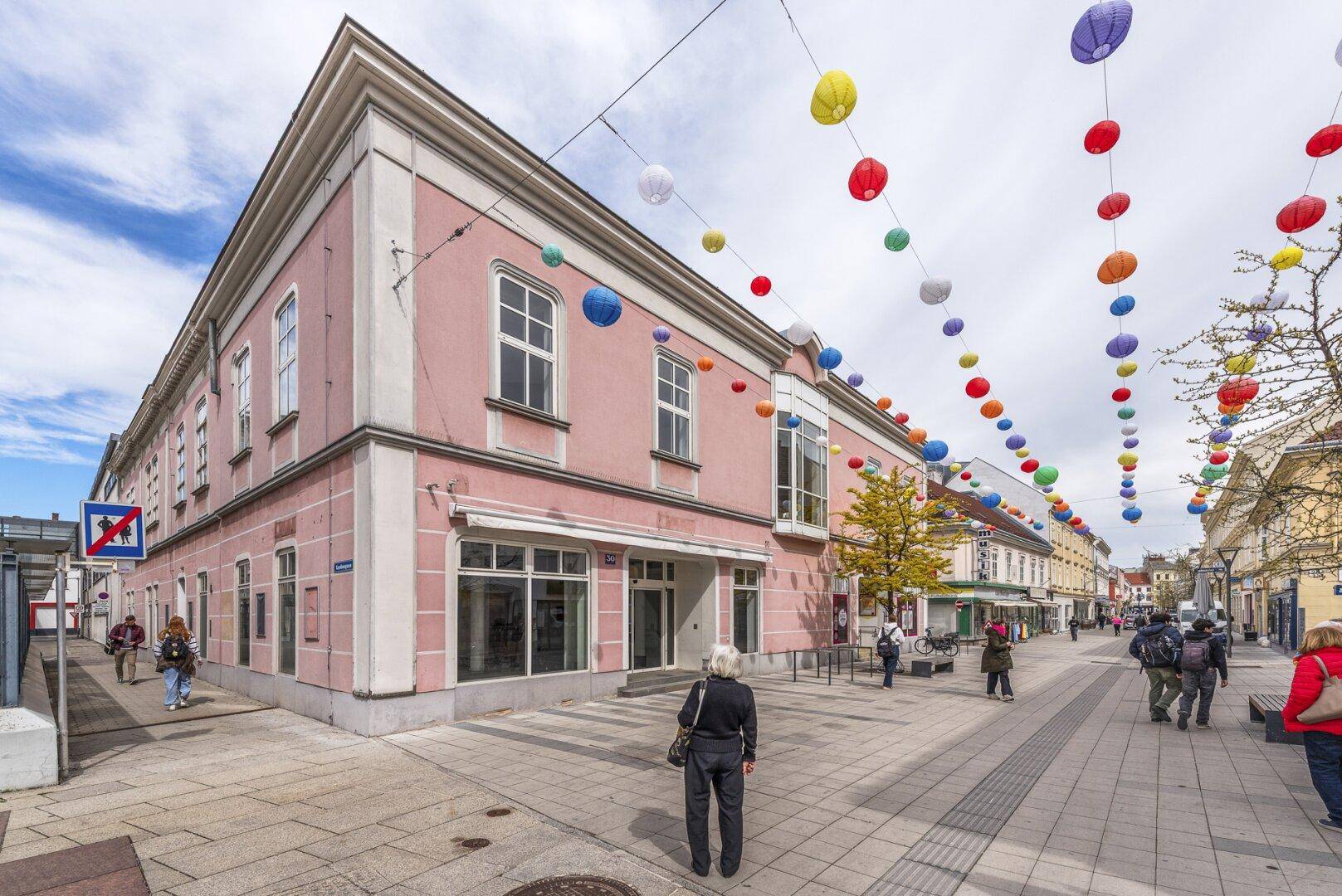 Eckgebäude mit rosa Fassade und großen Schaufenstern in einer belebten Fußgängerzone.