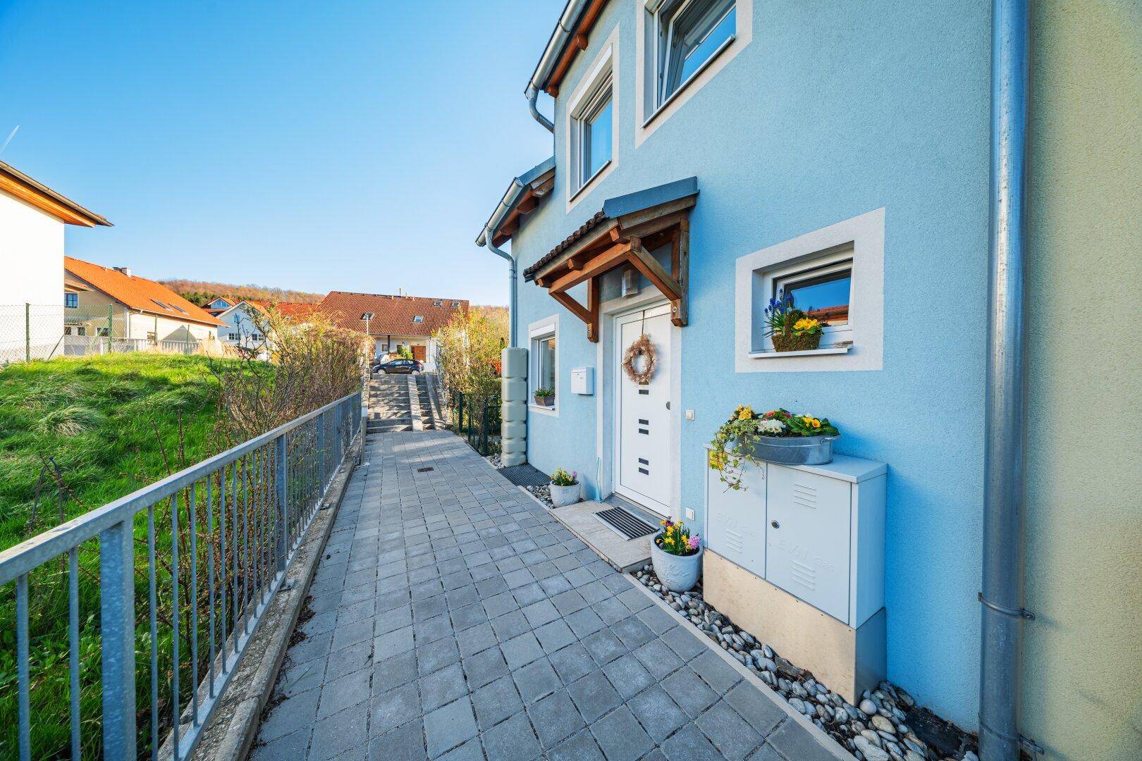 Vorderansicht des blauen Hauses mit gepflastertem Zugang, Haustür und dekorativen Blumenkästen.