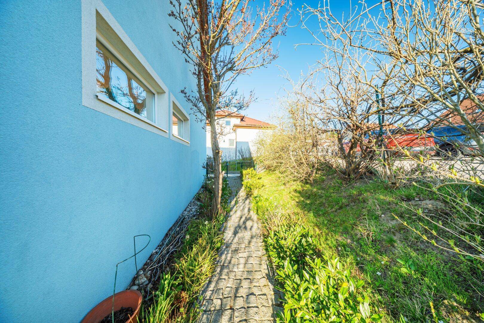 Schmaler gepflasterter Weg entlang der blauen Hausfassade mit grüner Bepflanzung und Fenstern.
