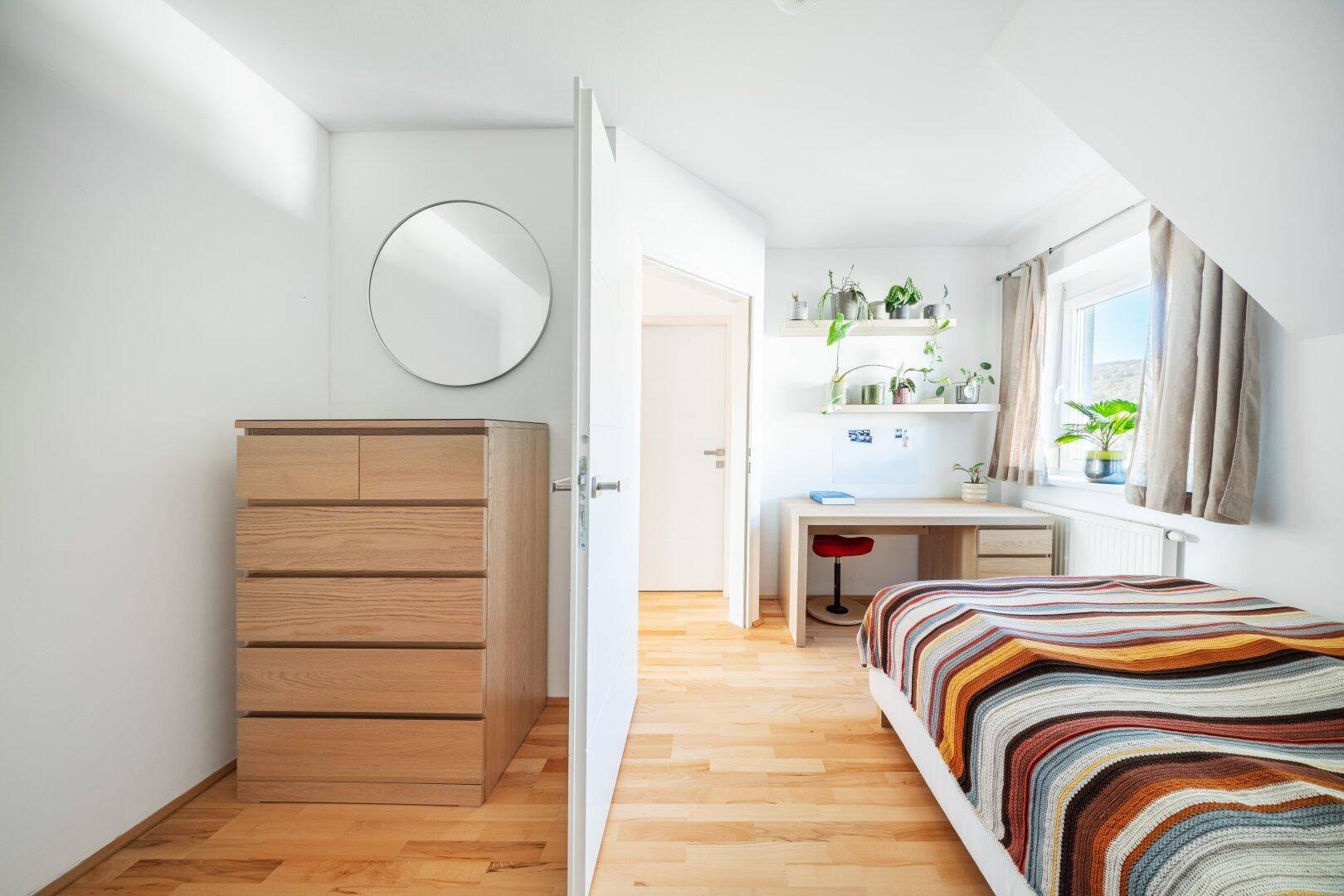 Helles Schlafzimmer mit Holzboden, Kommode und Schreibtisch am Fenster.