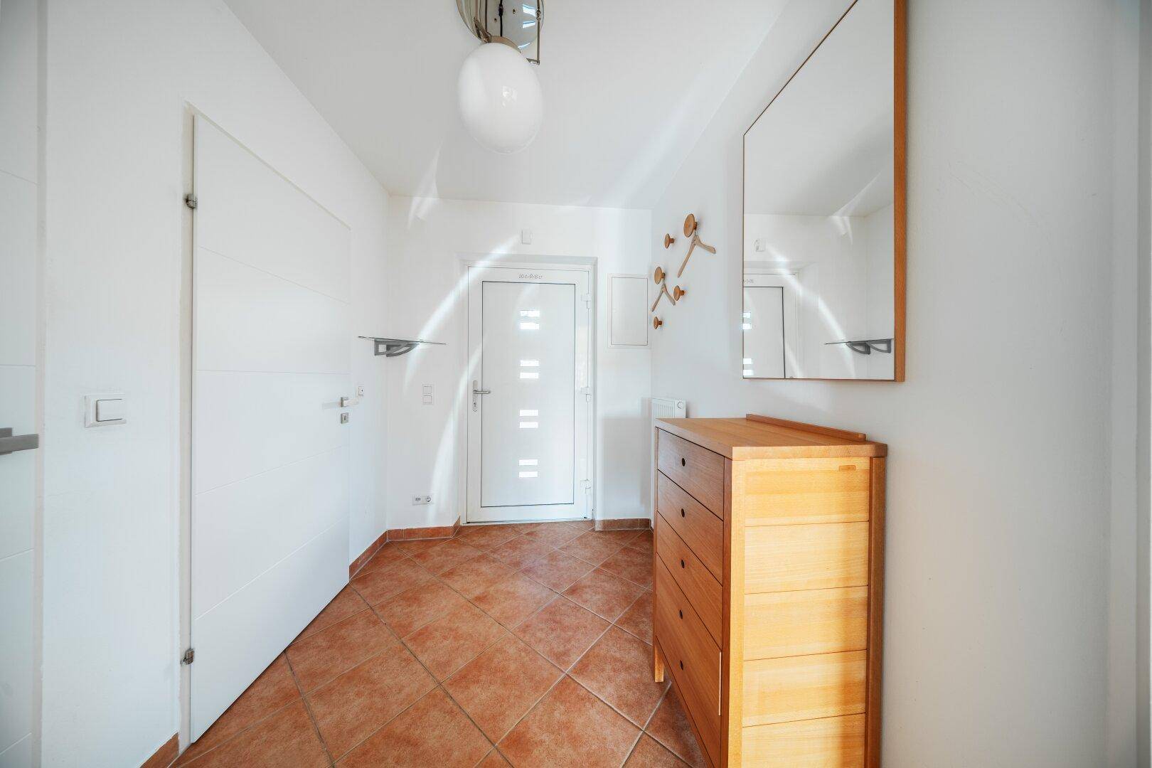 Eingangsbereich mit weißer Haustür, Holzkommode, Spiegel und praktischen Garderobenhaken.