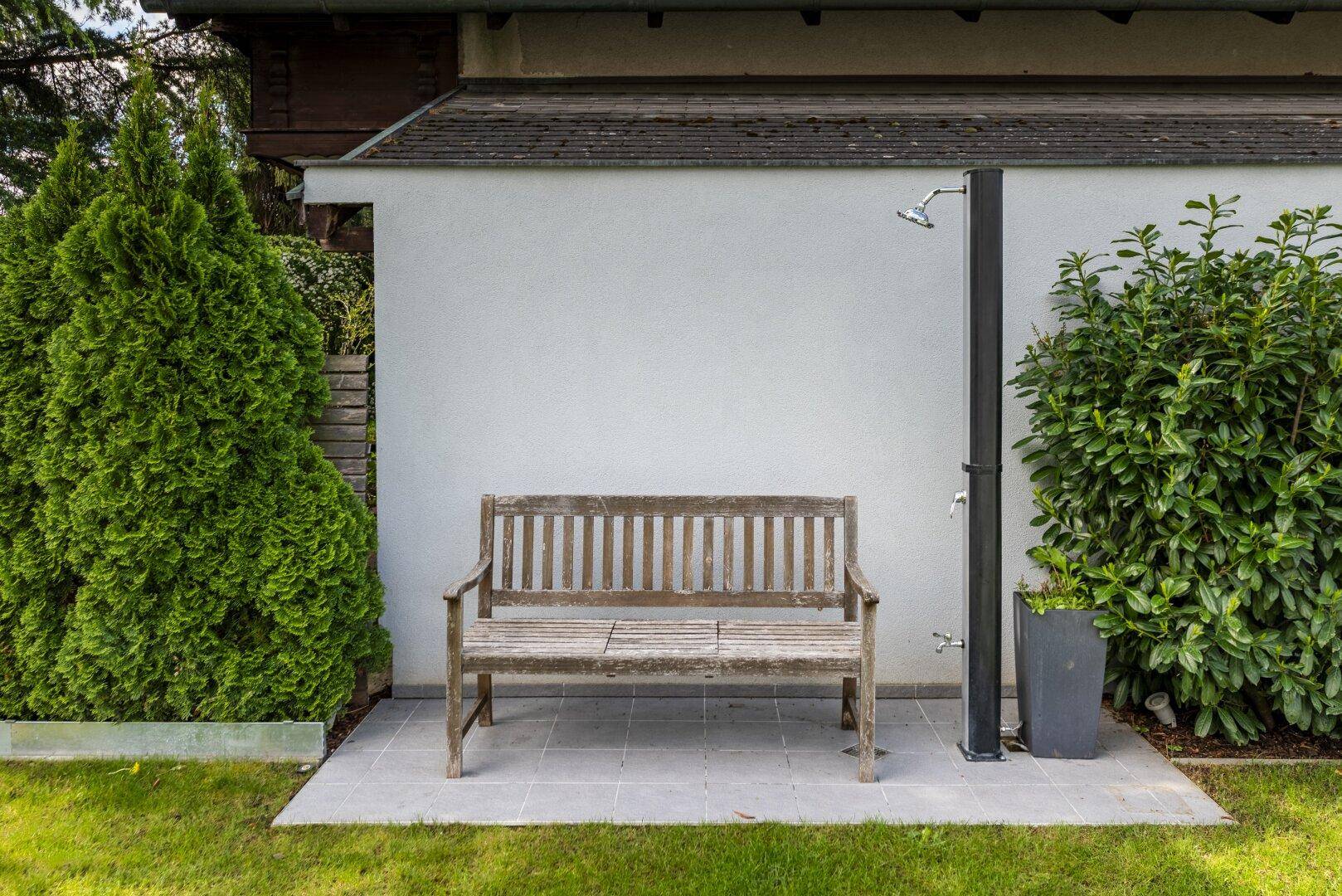 Gartenbereich mit Außendusche und Holzbank, umgeben von grünen Pflanzen.