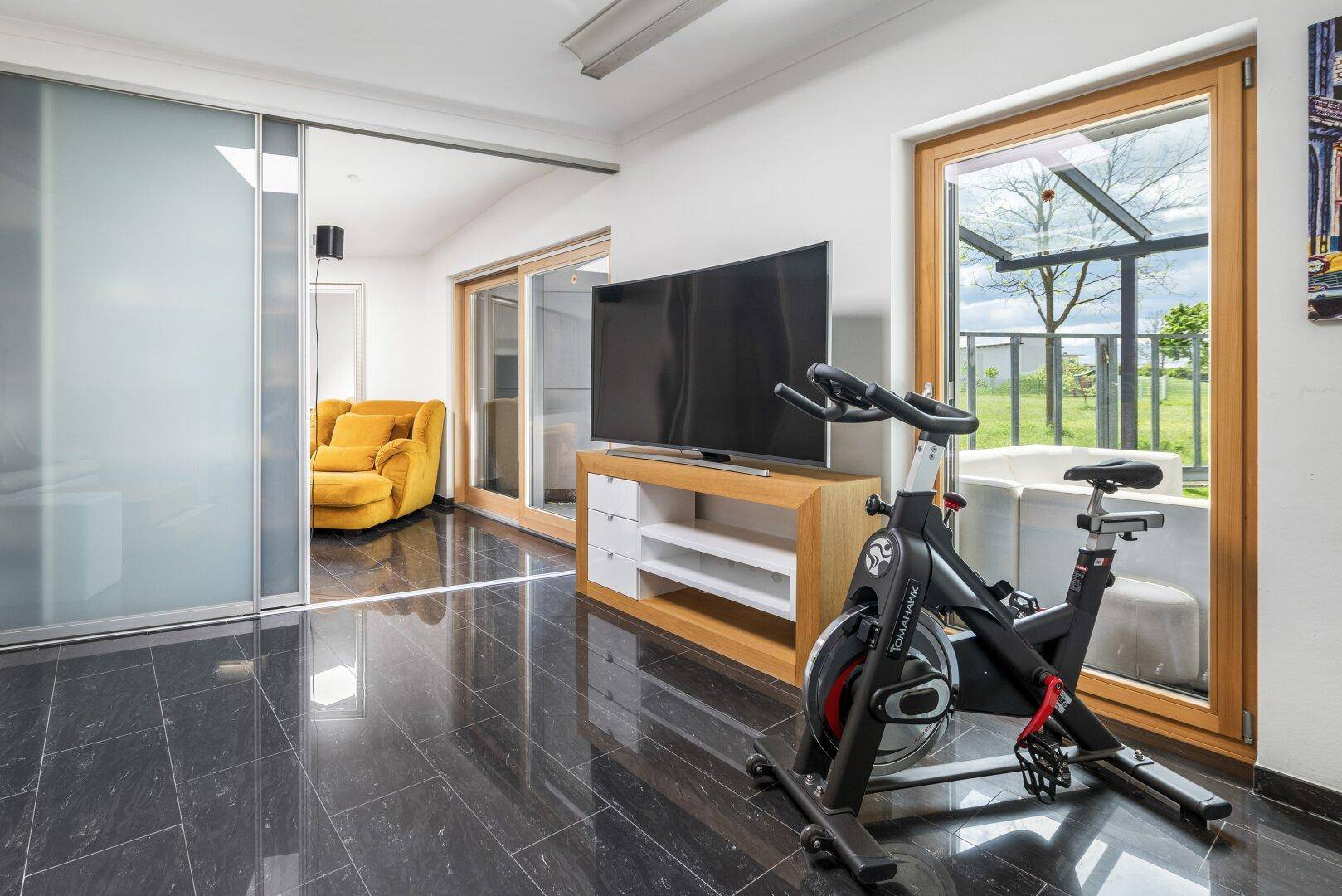 Fitnessraum mit Heimtrainer, großem Fernseher und Blick in einen Wintergarten oder Anbau.