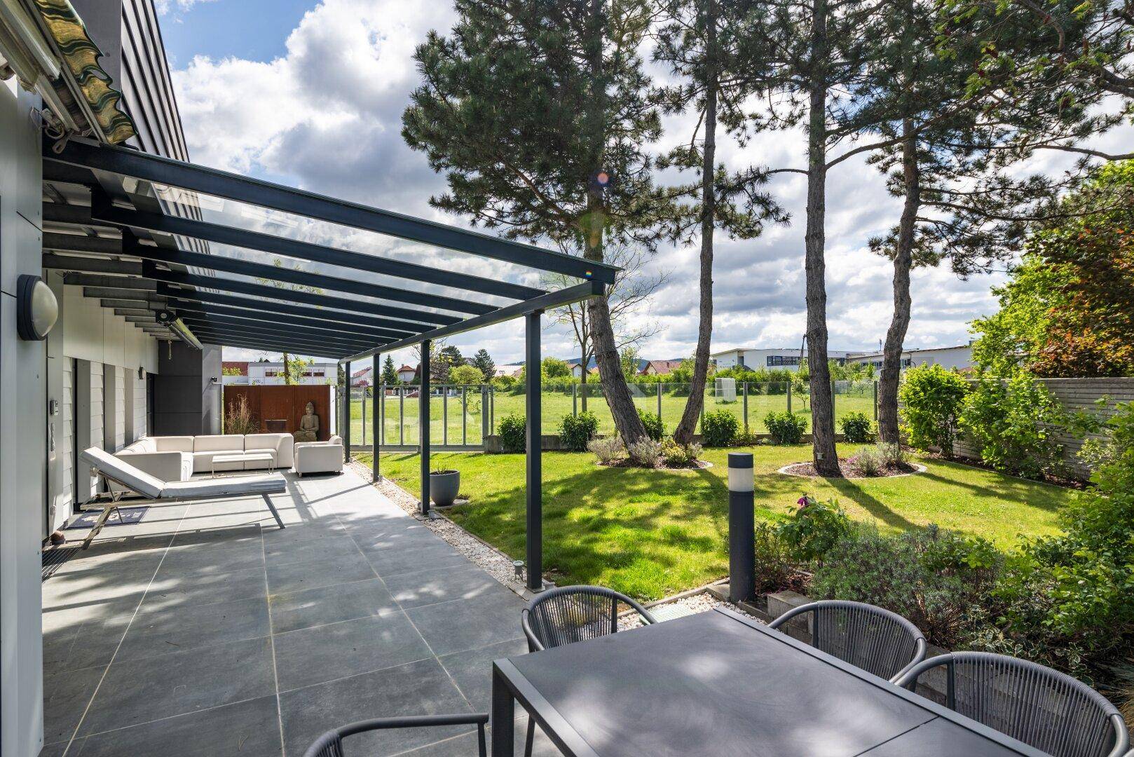 Überdachte Terrasse mit modernen Gartenmöbeln, angrenzendem Rasen und hohen Bäumen.