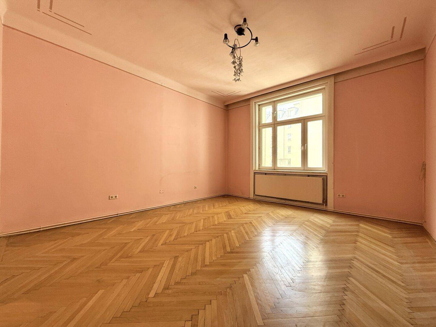 Geräumiges Zimmer mit rosa Wänden, Fischgrätparkett und einem großen Fenster.