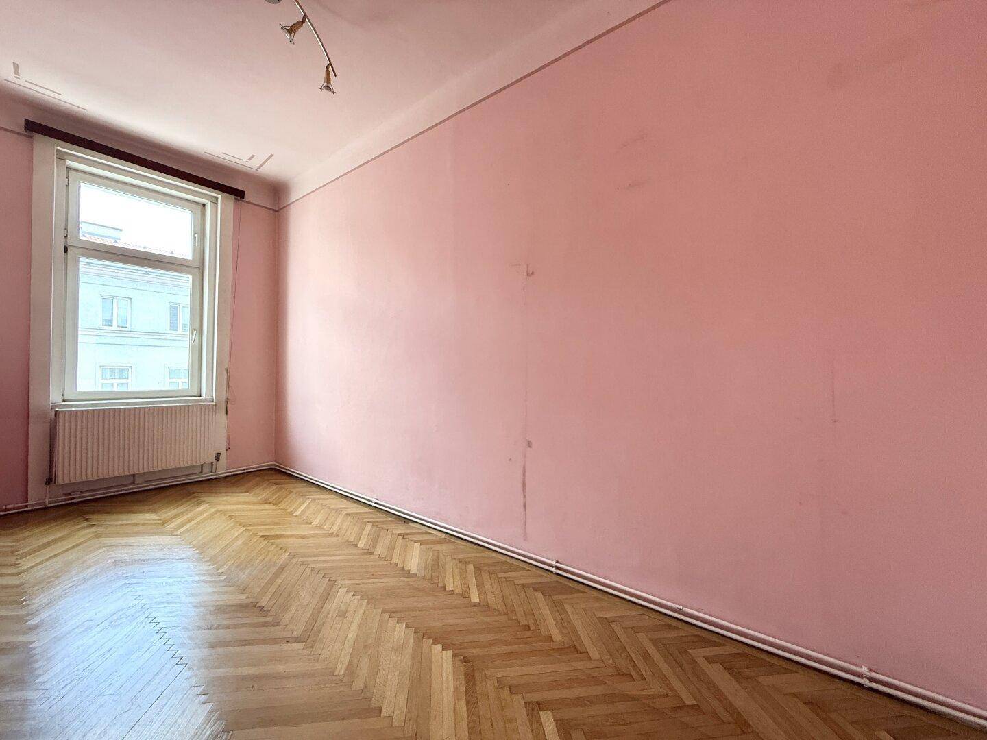 Leeres Zimmer mit auffälligen rosa Wänden, Fischgrätparkett und einem großen Fenster.