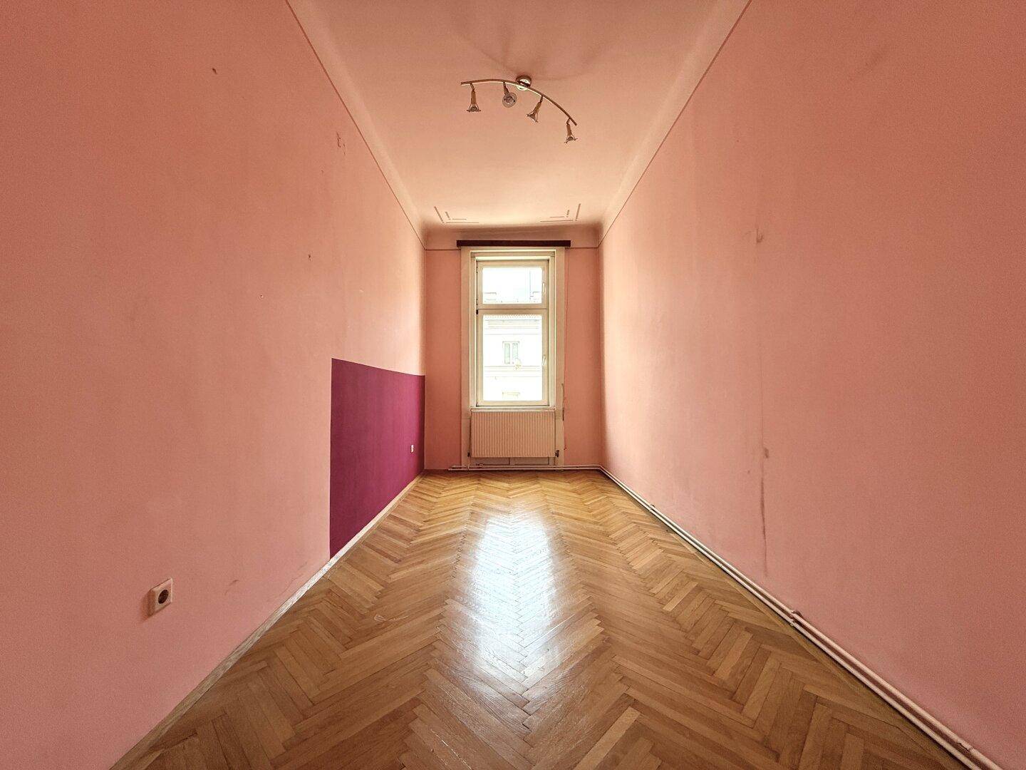Schmaler Raum mit rosa Wänden, Fischgrätparkett und einem Fenster am Ende des Zimmers.
