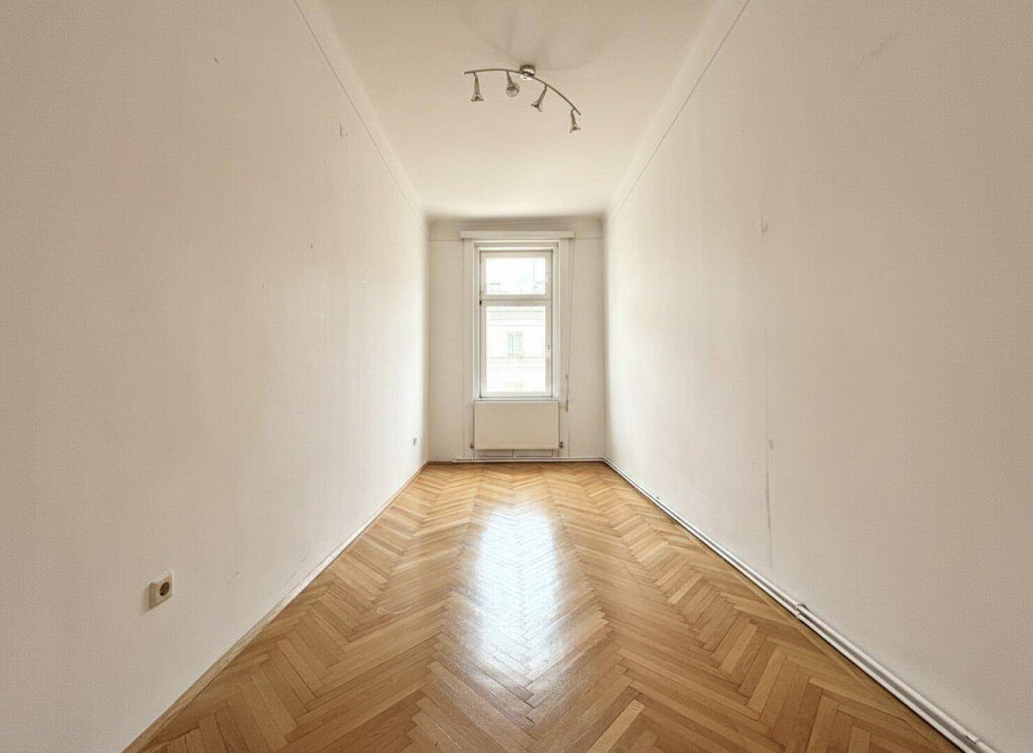 Schmaler, heller Raum mit weißen Wänden, Fischgrätparkett und einem Fenster am Ende.
