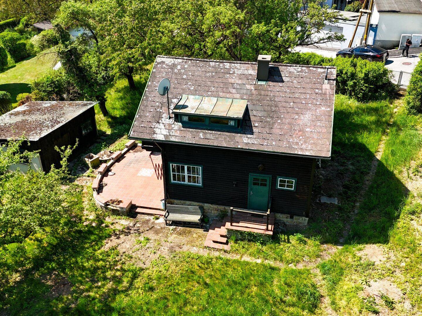 Vorderansicht des rustikalen Hauses mit kleiner Terrasse und Satteldach, umgeben von Grün.