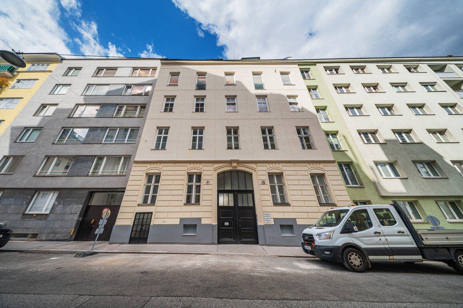 Fassade eines Mehrfamilienhauses an einer städtischen Straße mit parkenden Fahrzeugen.