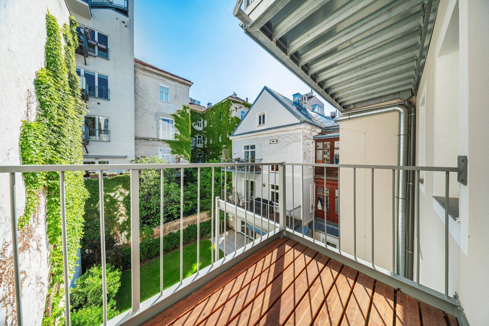 Kleiner Balkon mit Holzboden und Blick auf einen begrünten Innenhof und umliegende Gebäude.