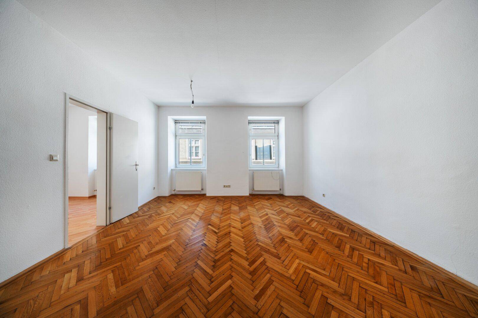 Geräumiges Zimmer mit klassischem Fischgrätparkett und zwei großen Fenstern.