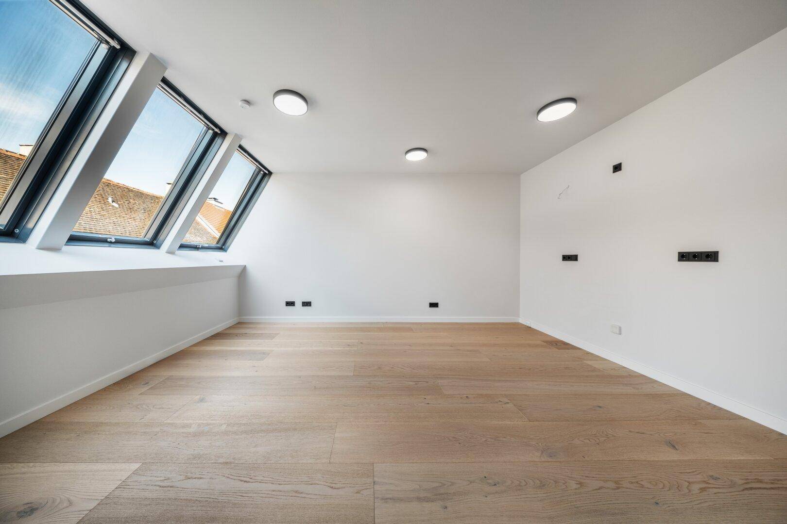 Helles Dachzimmer mit Holzboden, schrägen Wänden und mehreren Dachfenstern für viel Licht.