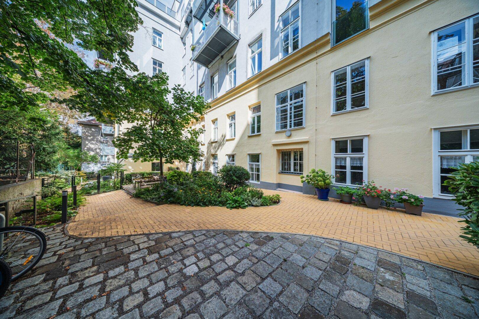 Gepflegter Innenhof mit Gartenbereich, Bäumen und gepflasterten Wegen vor einer gelben Gebäudefassade.