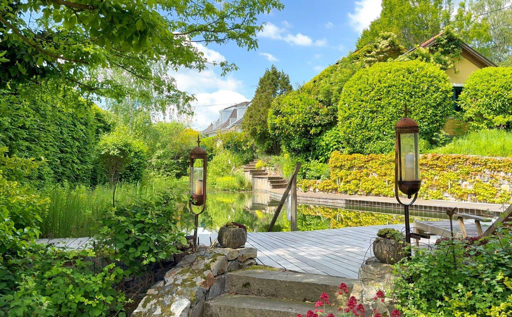 Üppiger grüner Garten mit Naturpool, Holzdeck und Steintreppen unter blauem Himmel.
