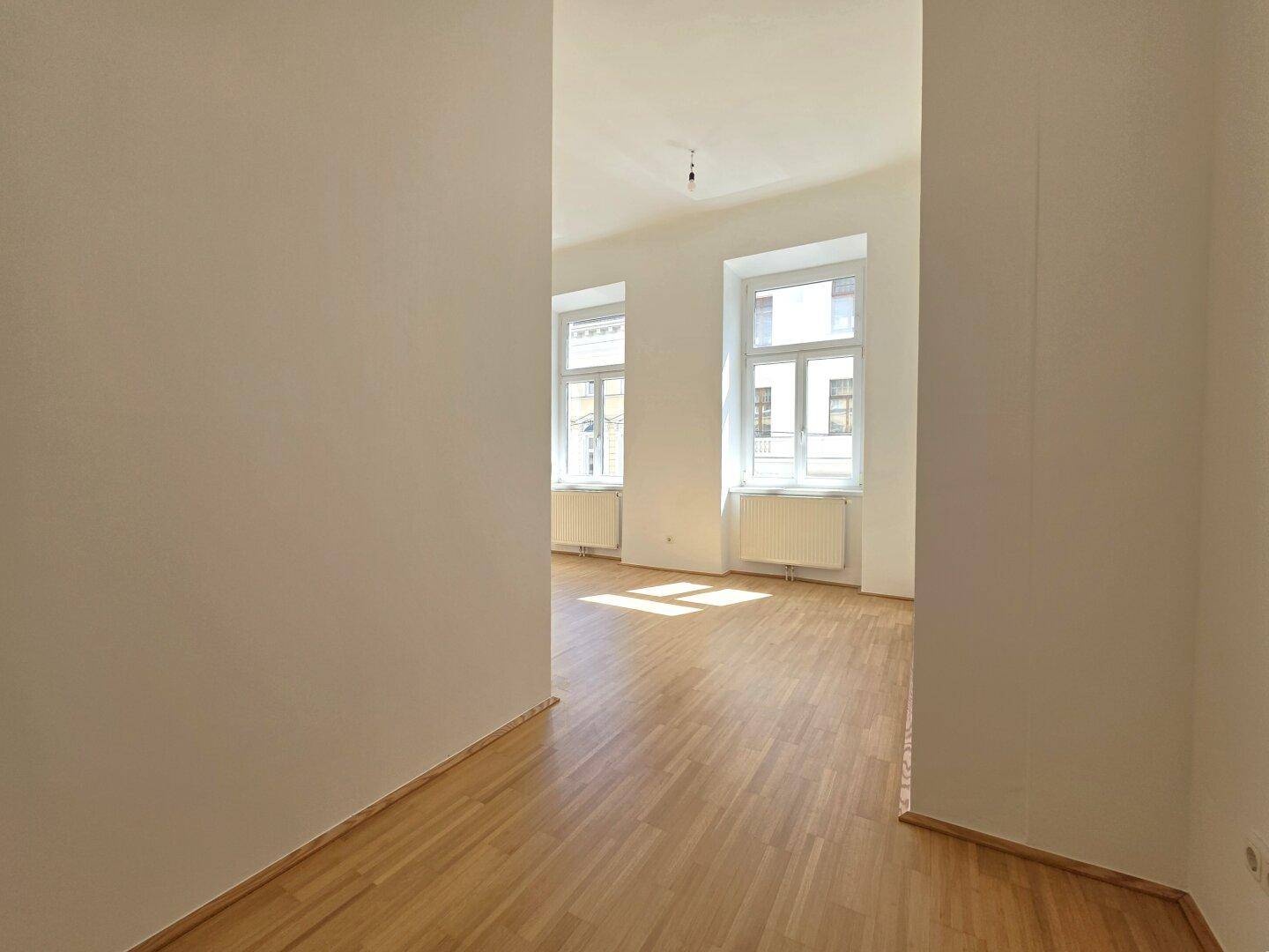 Heller Flur mit Parkettboden, der in einen lichtdurchfluteten Raum mit Fenstern führt.