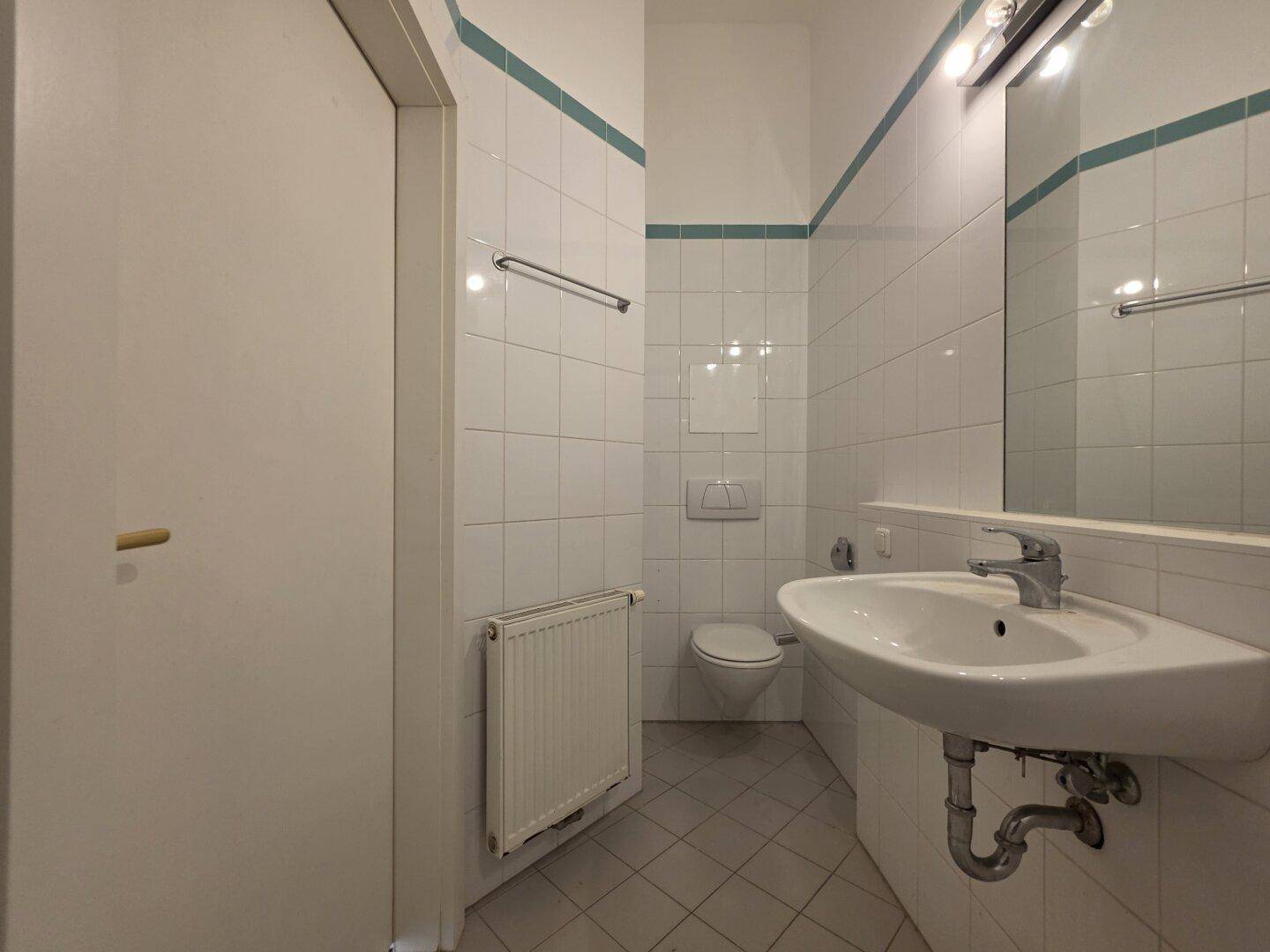 Helles Badezimmer mit weißen Fliesen, Waschbecken und Toilette, ausgestattet mit Heizkörper.
