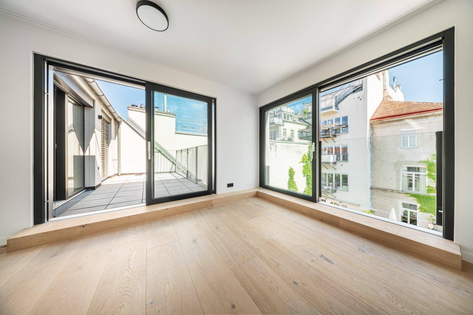 Helles Zimmer mit Holzboden, großen Fenstern und Zugang zu einem Außenbereich mit Hofblick.