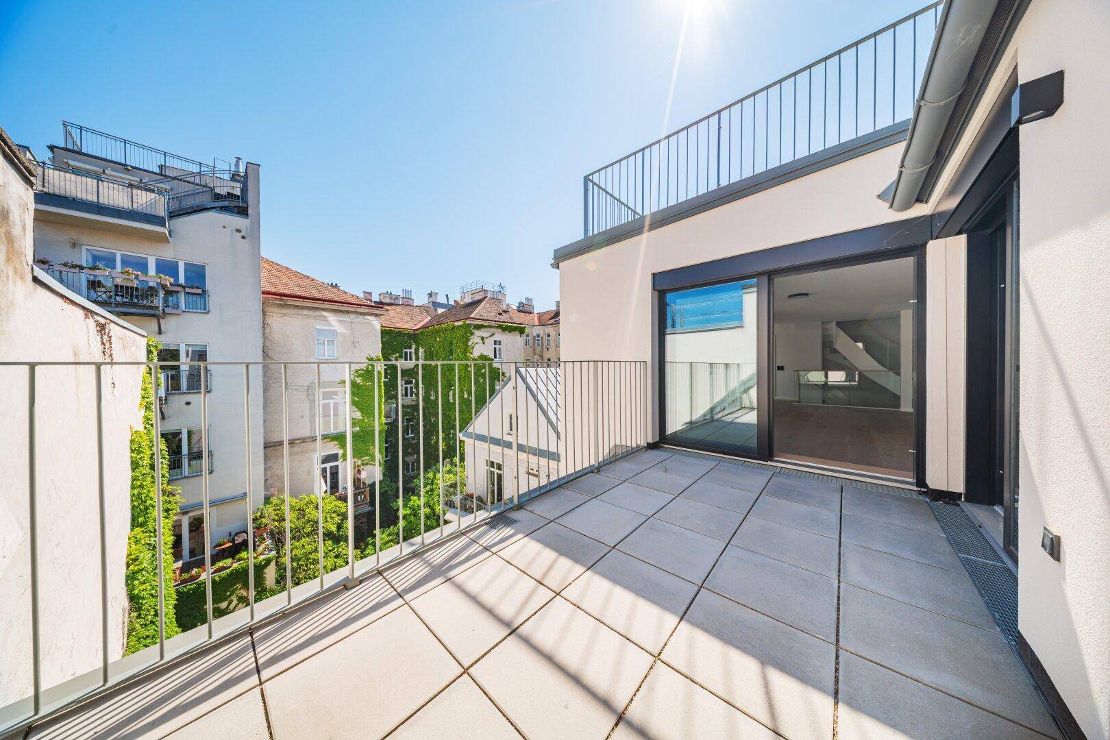 Heller Balkon mit Fliesenboden und Blick auf einen begrünten Innenhof und umliegende Gebäude.
