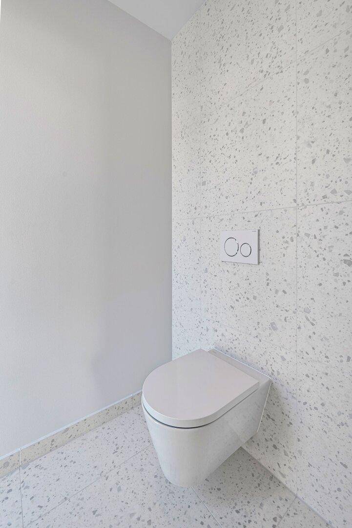 Modernes WC mit wandhängender Toilette und hellgrau gesprenkelten Fliesen an Boden und Wand.