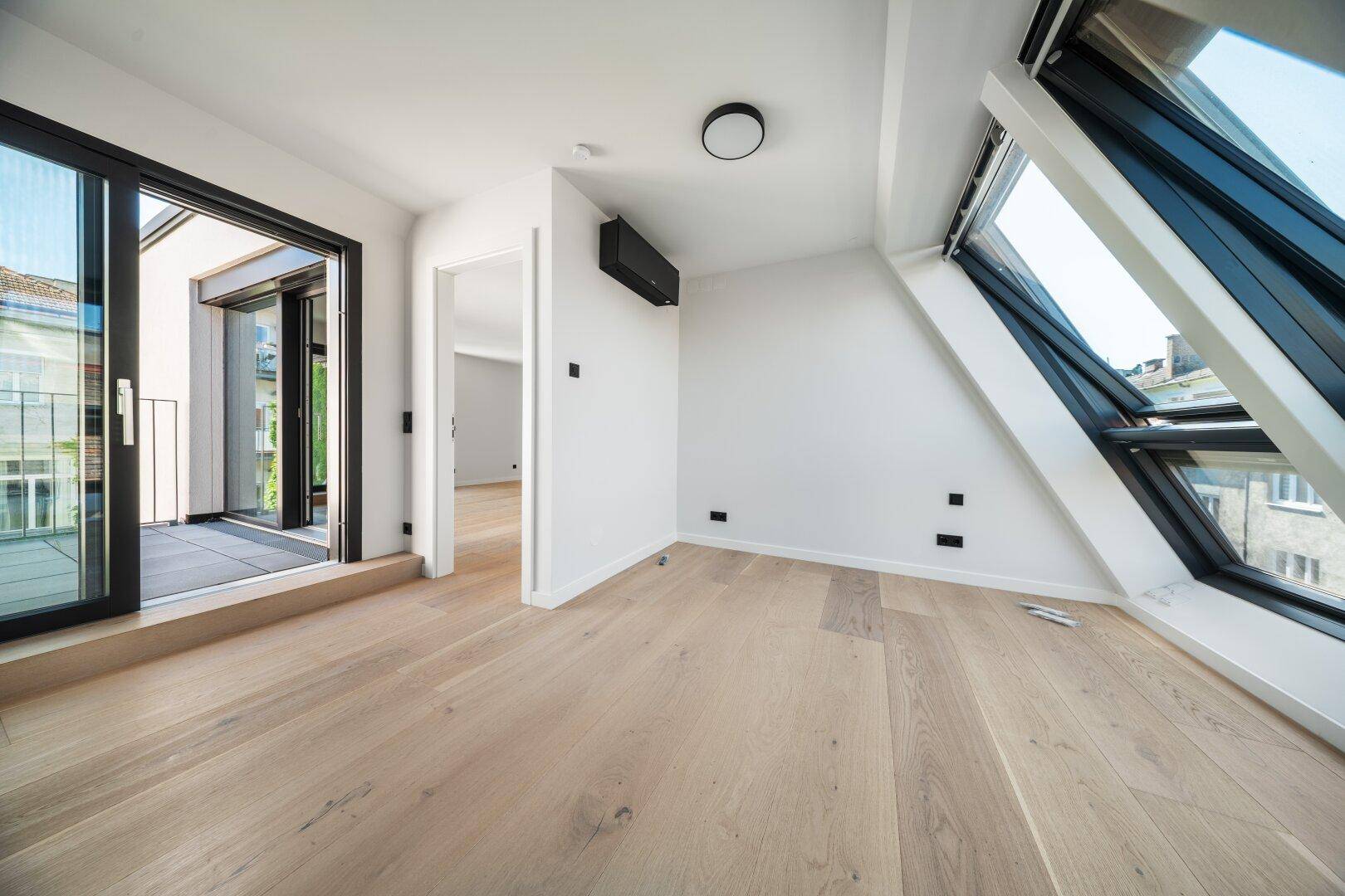 Lichtdurchflutetes Dachgeschosszimmer mit Holzboden, Klimaanlage und Zugang zu einem Balkon.