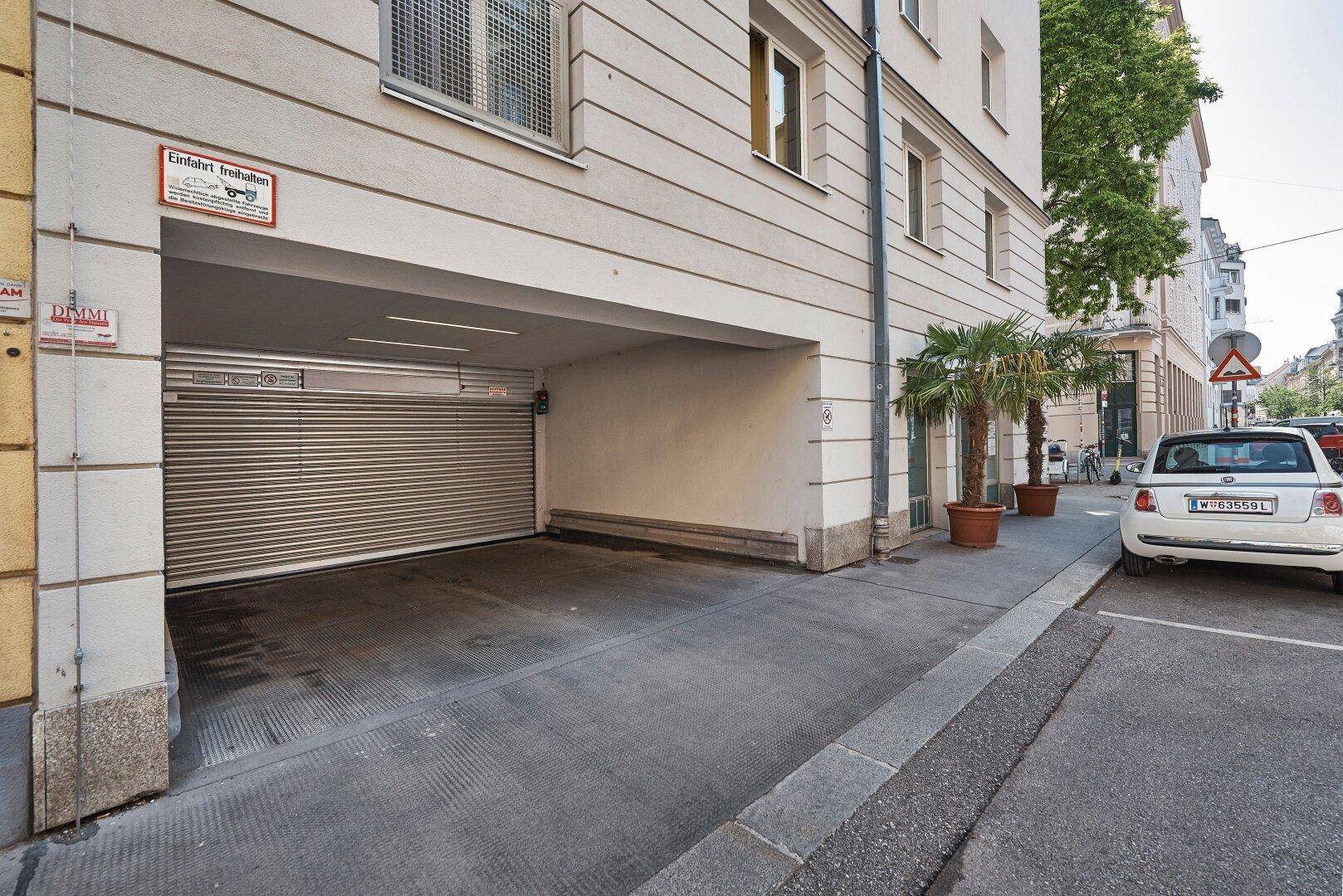 Einfahrt zu einer Tiefgarage mit Rolltor, flankiert von Palmen in Töpfen und einem geparkten Auto.