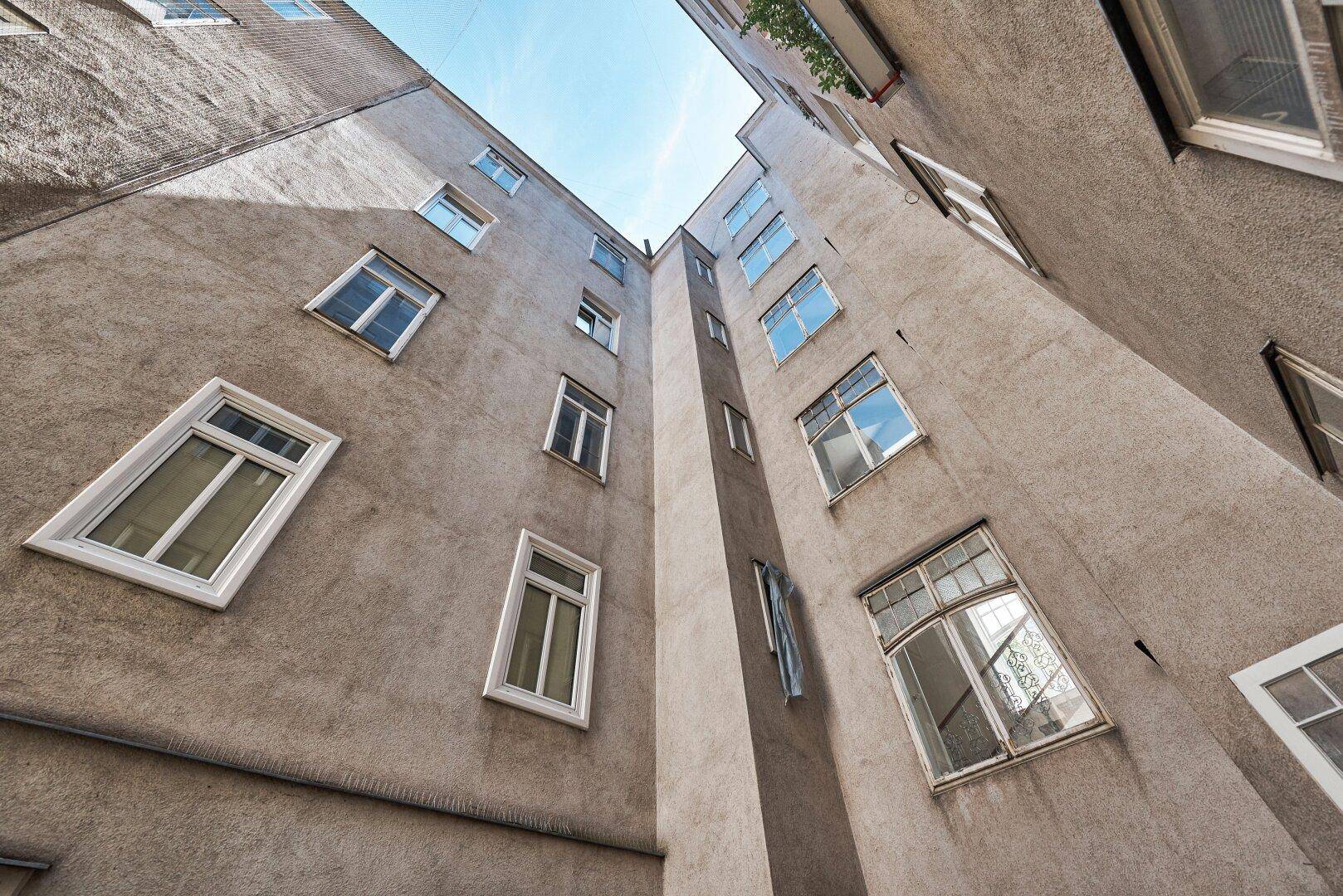 Blick nach oben in einen engen Innenhof eines Altbaus mit vielen Fenstern und strukturierter Fassade.