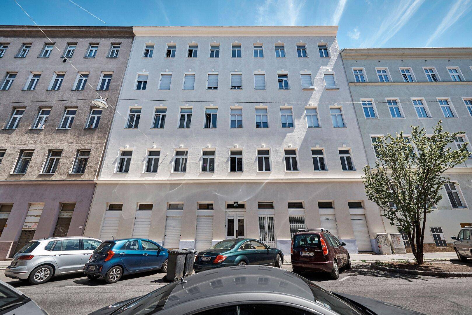 Straßenansicht eines Wohngebäudes mit hellblauer Fassade und parkenden Autos davor an einem sonnigen Tag.