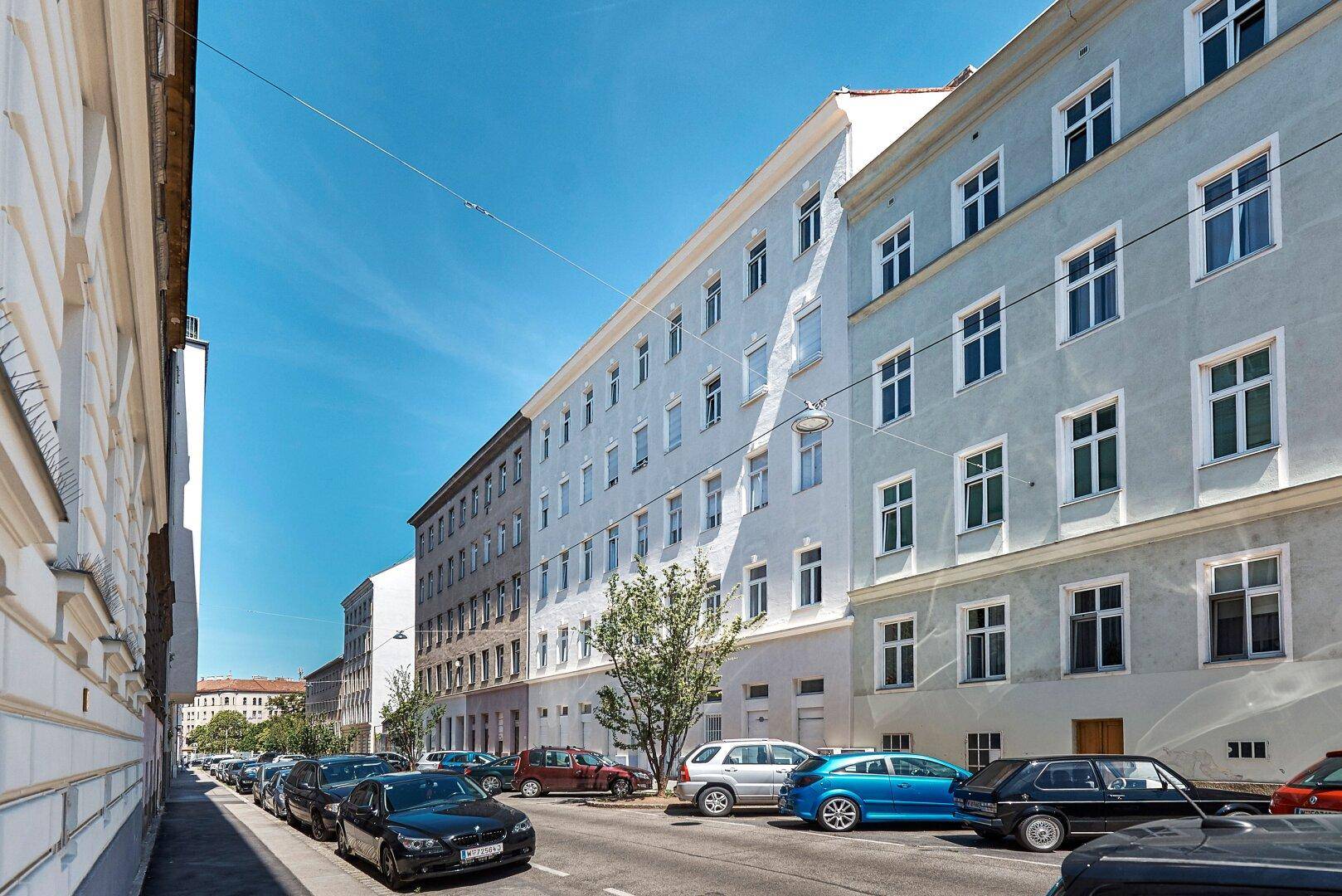 Weitläufige Straßenansicht mit mehreren Wohngebäuden und parkenden Autos unter blauem Himmel.