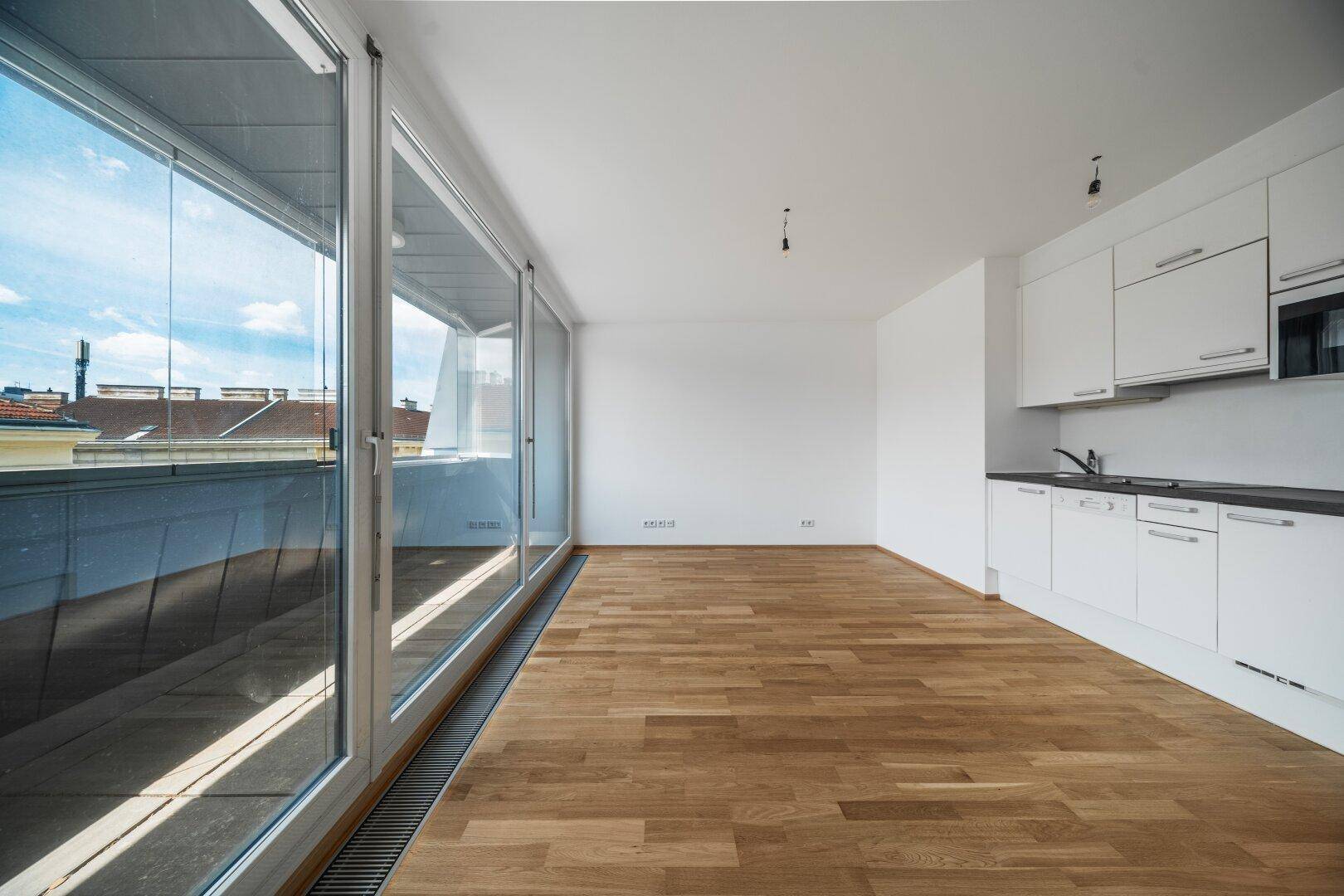 Offener Wohnbereich mit integrierter Küchenzeile, Holzboden und Zugang zum verglasten Balkon.