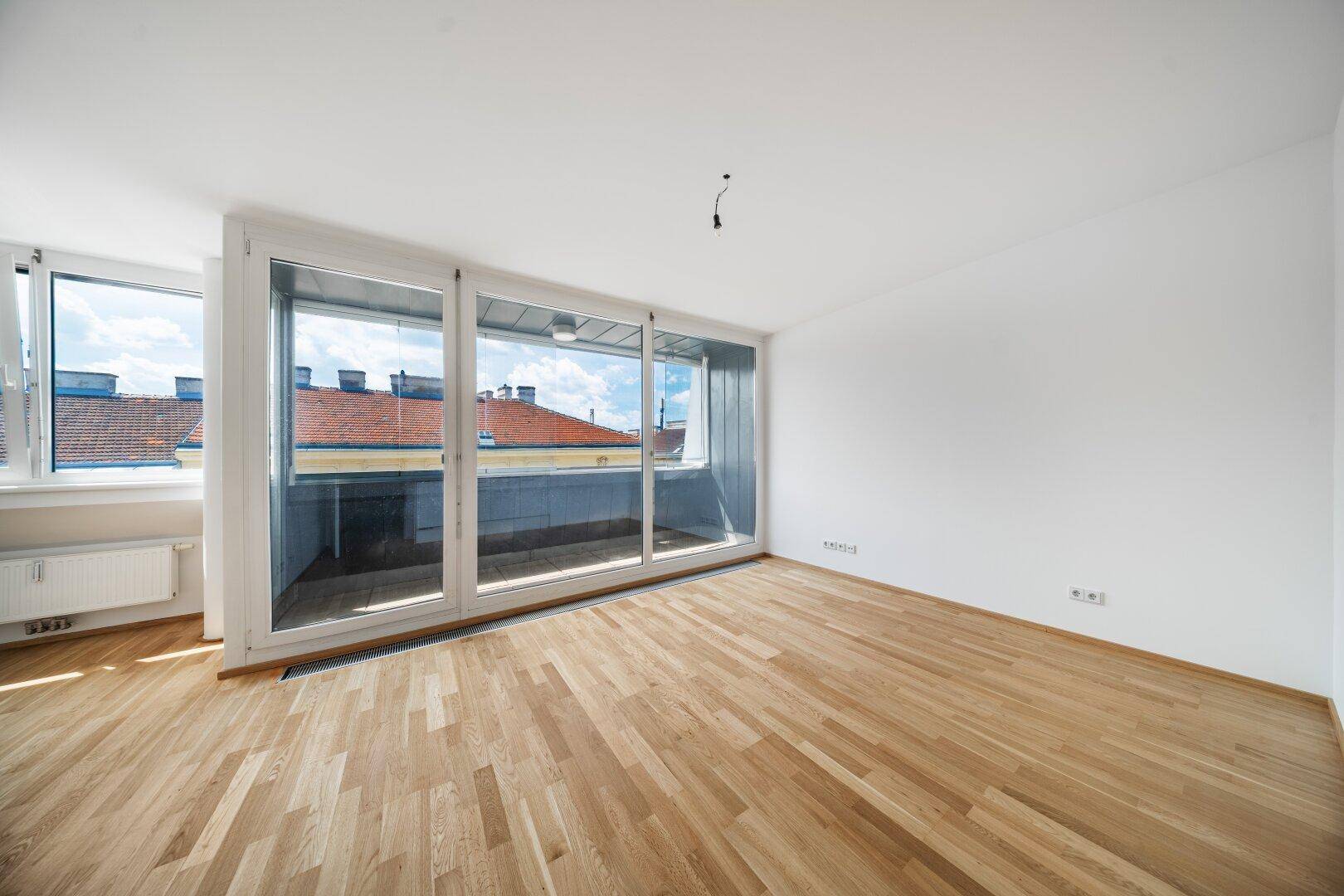 Geräumiges Zimmer mit hellem Holzboden und großen Fenstern, die viel Tageslicht hereinlassen.