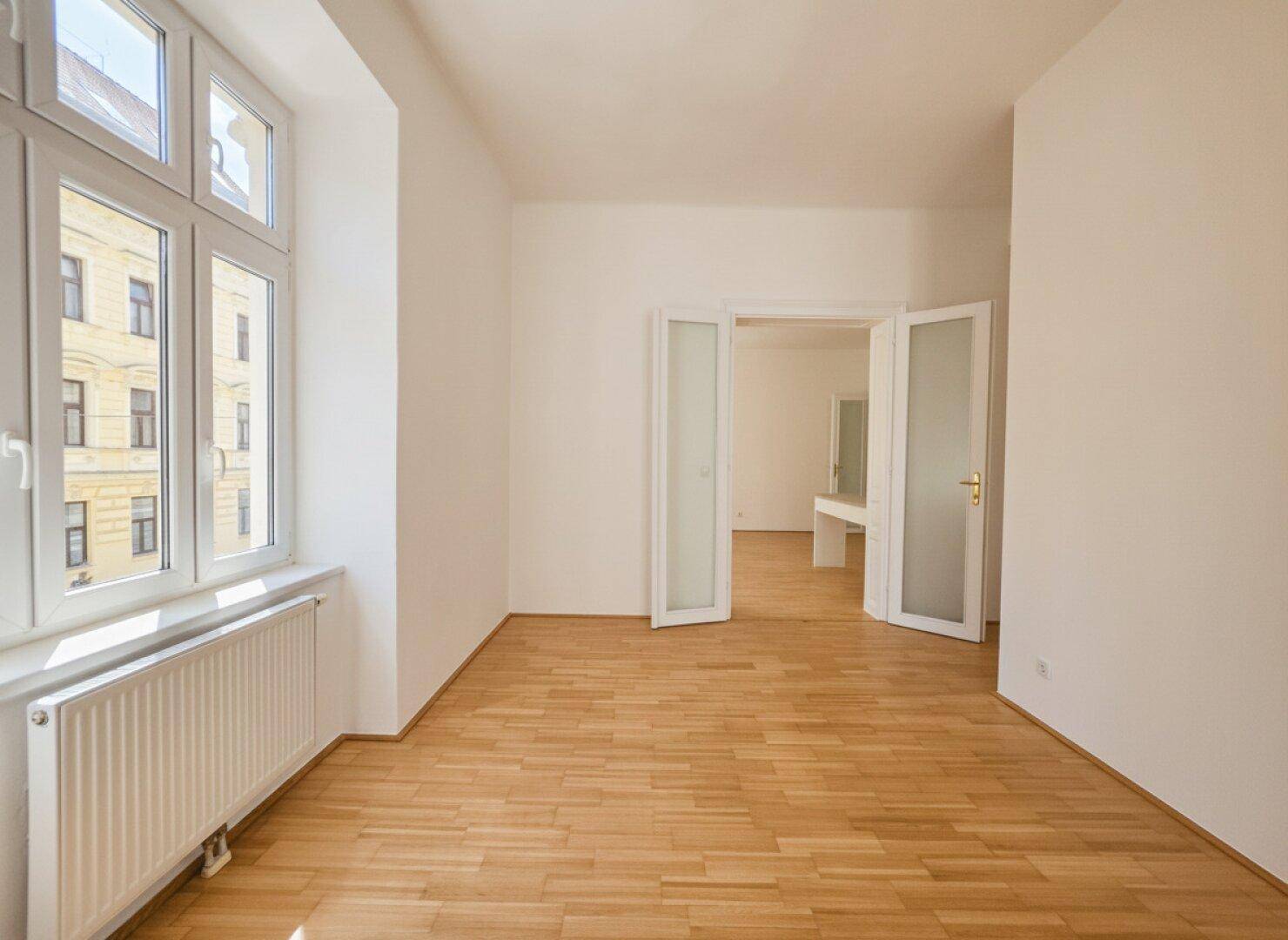 Heller Wohnraum mit Parkettboden und eleganten Doppeltüren, die zu einem weiteren Zimmer führen.