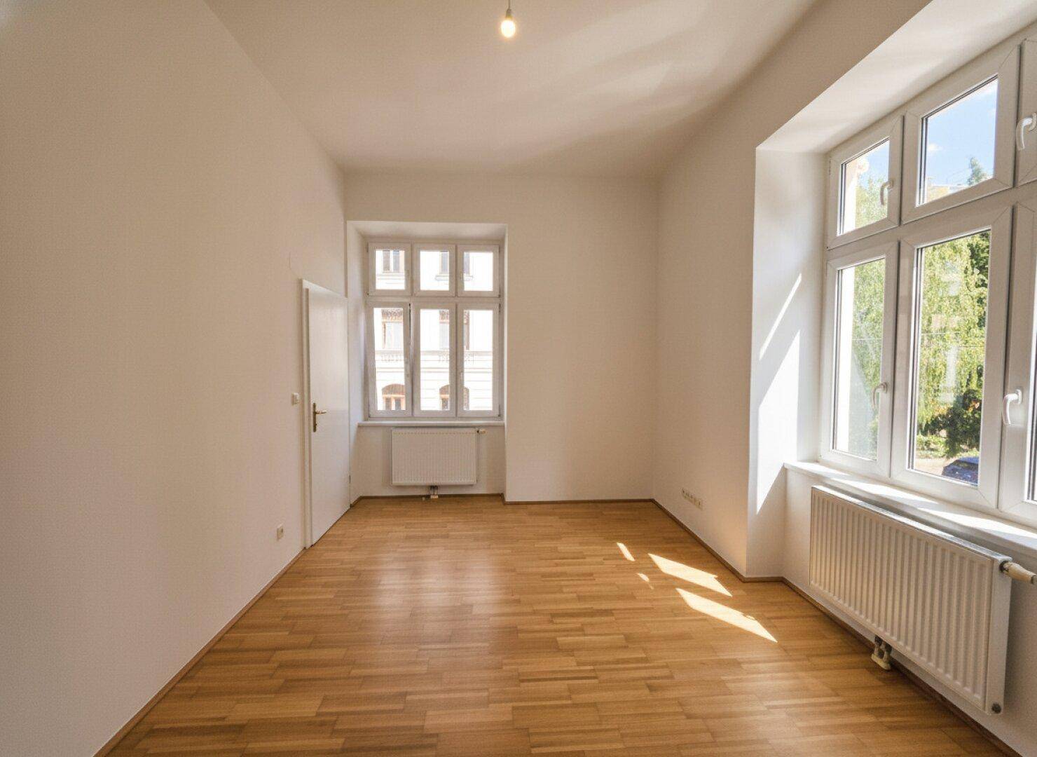 Helles Zimmer mit Parkettboden und großen Fenstern, die einen Blick ins Grüne ermöglichen.