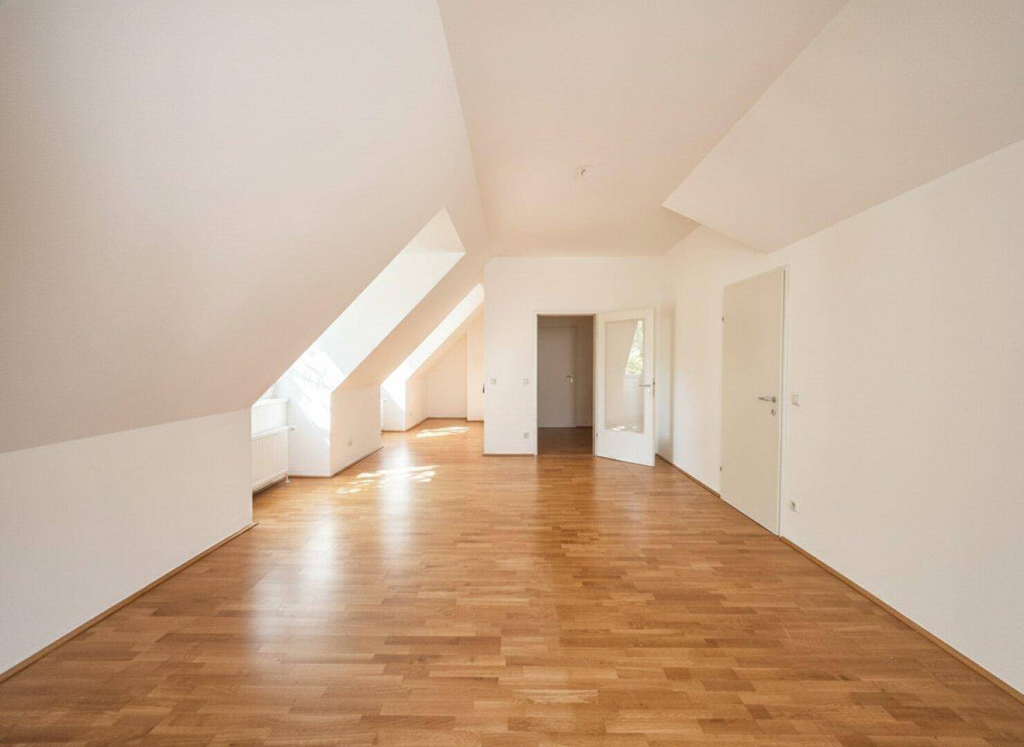 Geräumiger Dachgeschossraum mit Holzboden, schrägen Wänden und offener Tür zu einem weiteren Bereich.