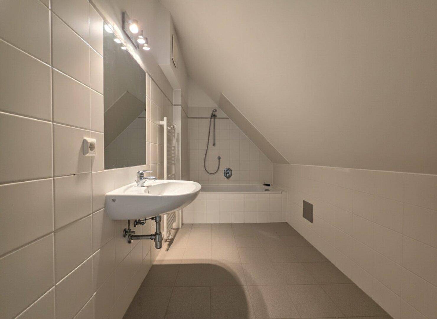 Badezimmer im Dachgeschoss mit Badewanne, Waschbecken, Spiegel und weißen Fliesen unter schräger Decke.