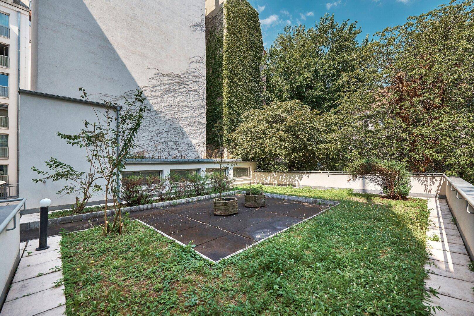 Begrünter Innenhof oder Gartenbereich mit Bäumen und Pflanzen, umgeben von Gebäudefassaden.