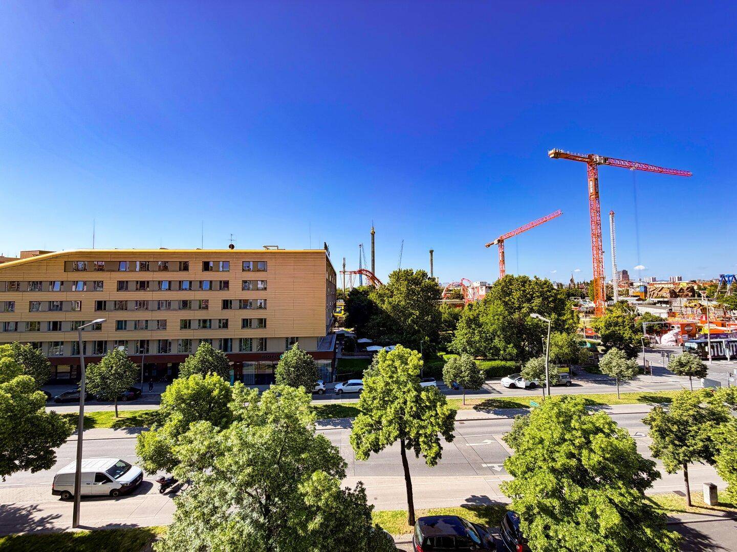 Weitblick über die Stadt mit einem modernen Gebäude, viel Grün und Baukränen am Horizont unter blauem Himmel.