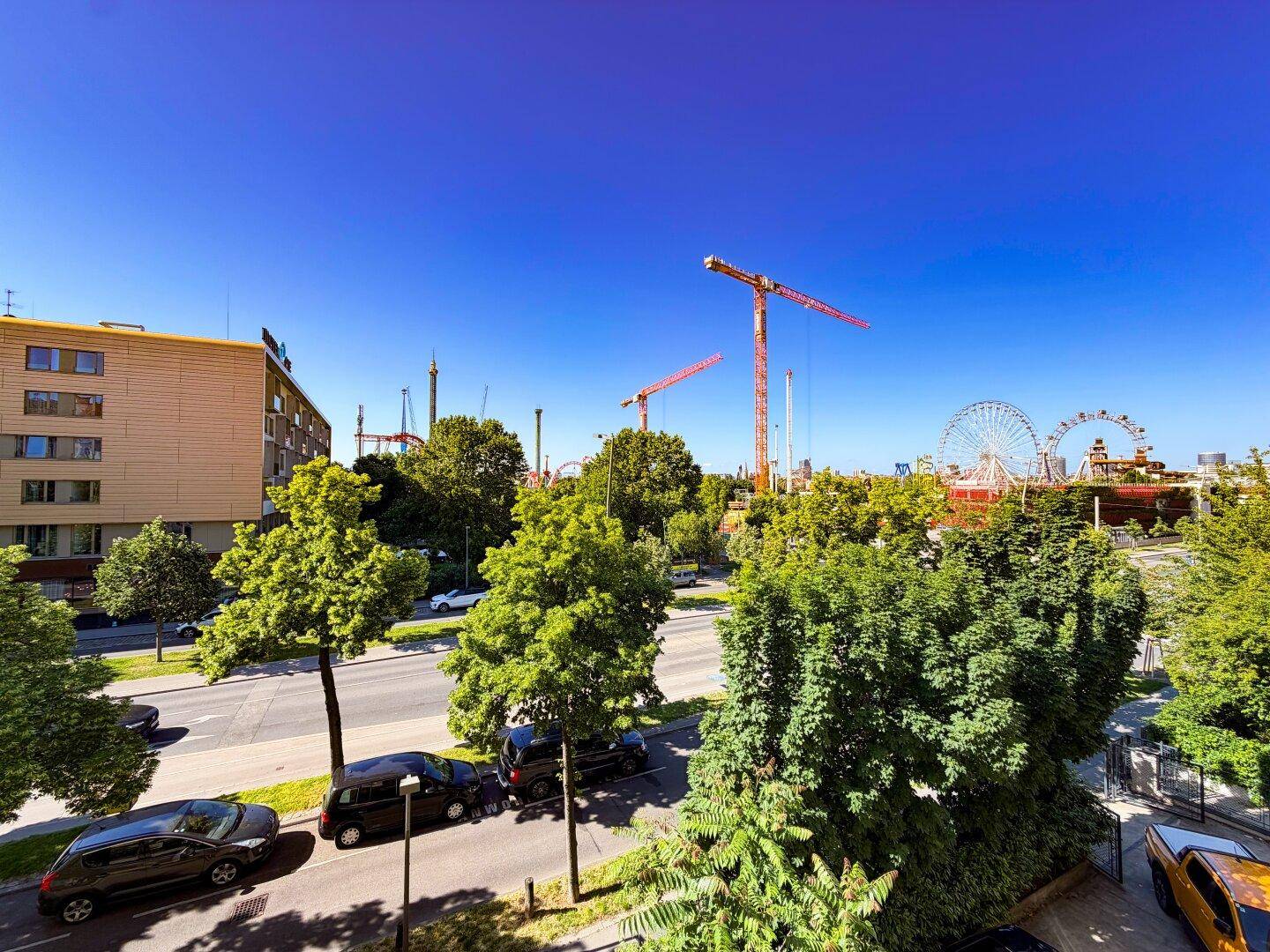 Blick auf die belebte Straße, grüne Bäume und den Prater mit Riesenrad und Achterbahnen.