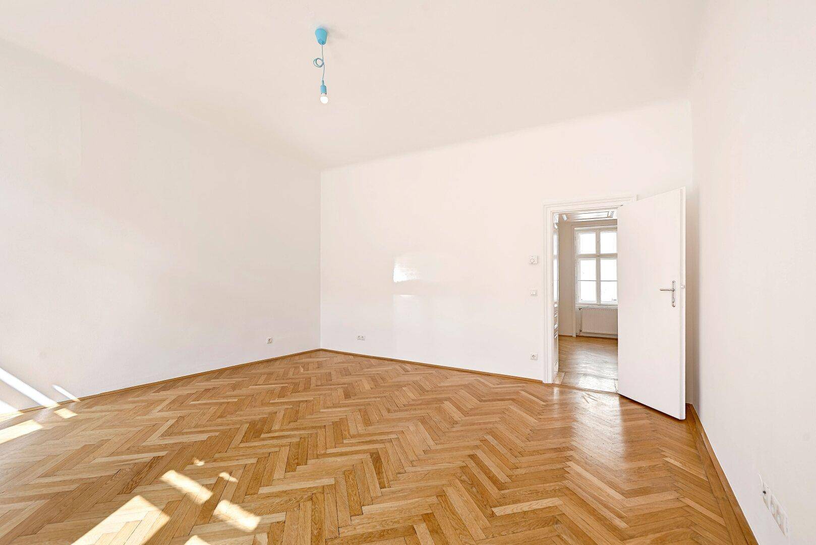 Geräumiges, helles Zimmer mit Fischgrätparkett und Zugang zu einem weiteren Raum.