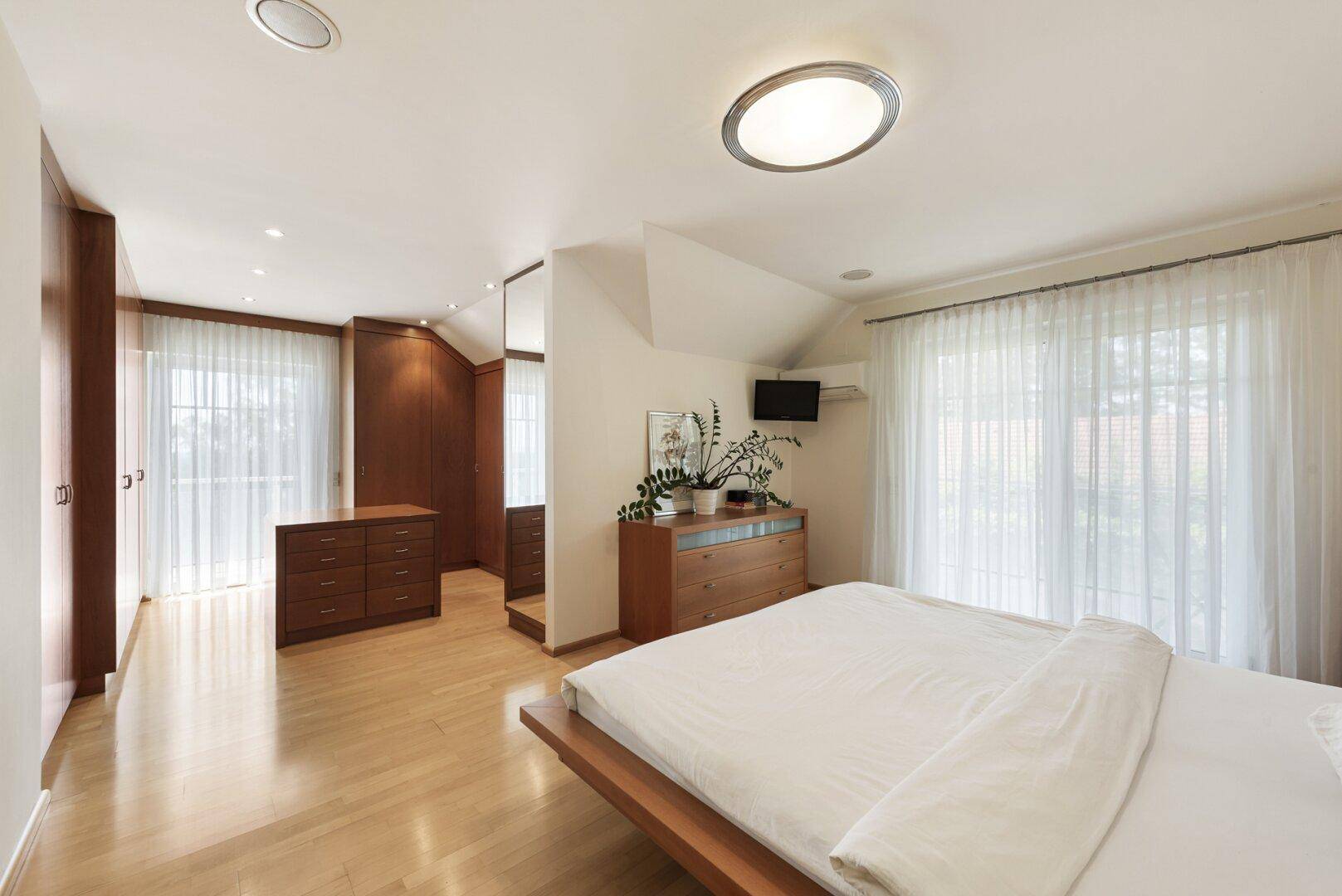 Geräumiges Schlafzimmer mit Holzboden, Einbauschränken und großen Fenstern mit Vorhängen.