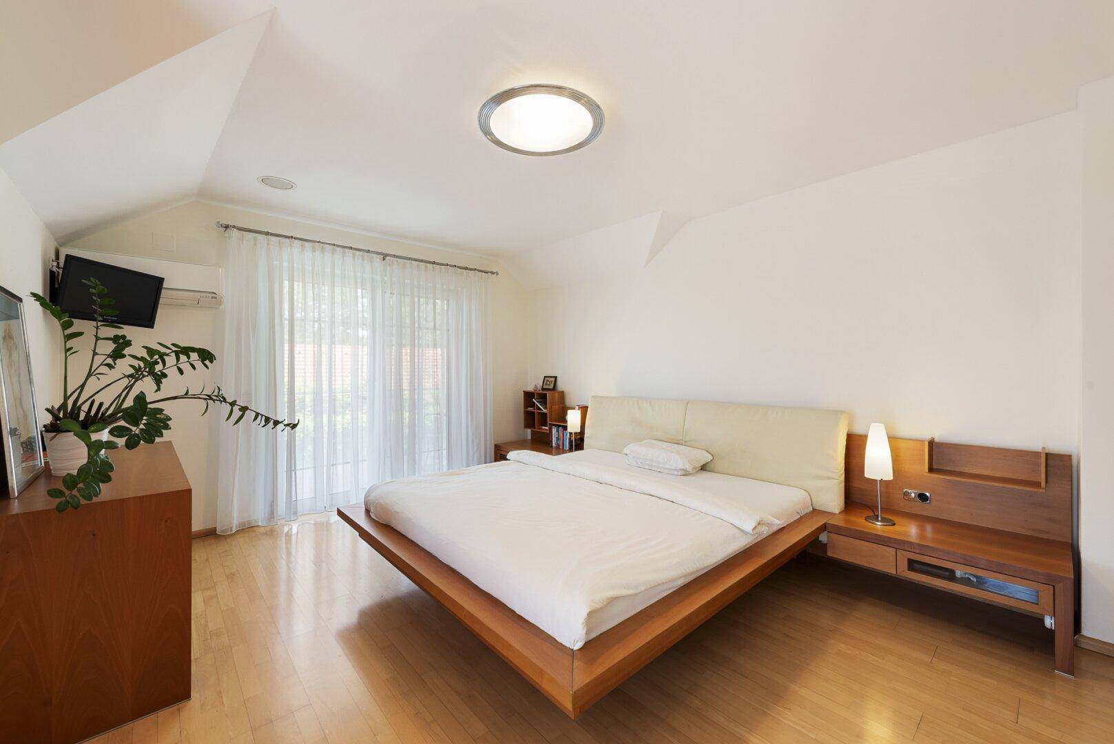 Helles Schlafzimmer mit Holzbett, Nachttischen und großen Fenstern mit Blick ins Grüne.