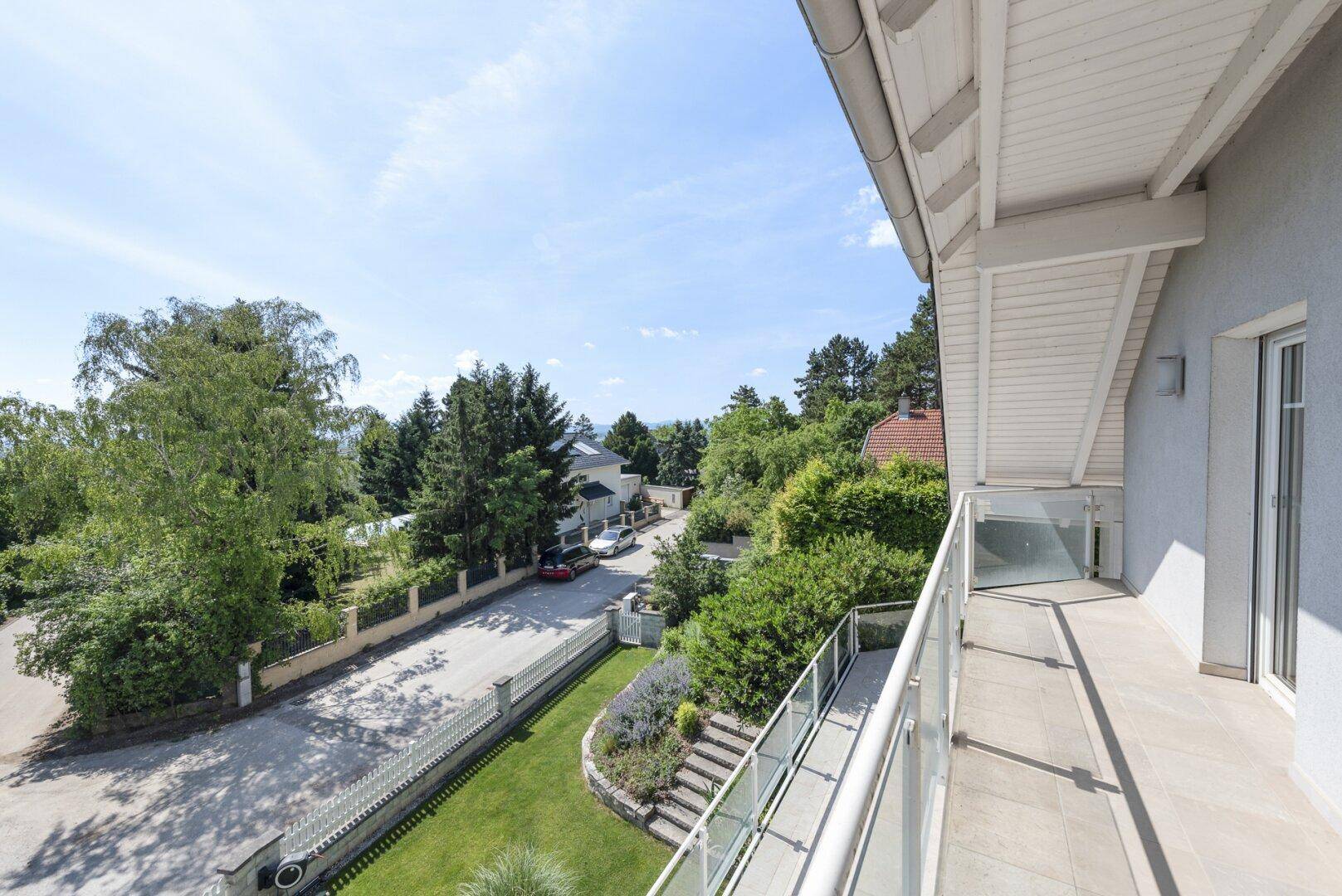 Balkon mit Glasgeländer und weitem Blick über die grüne Umgebung und die Straße.