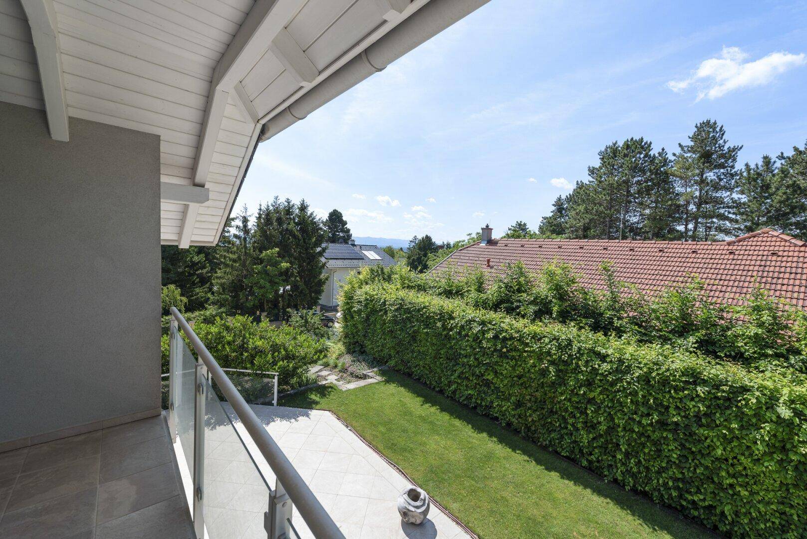 Balkon mit Glasgeländer und weitem Blick über den grünen Garten und die Umgebung.