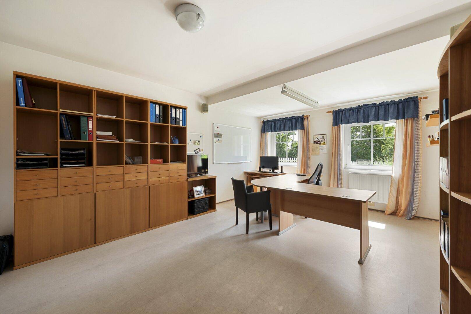 Großes Arbeitszimmer mit Holzregalen, Schreibtisch und Fenstern mit Blick ins Grüne.