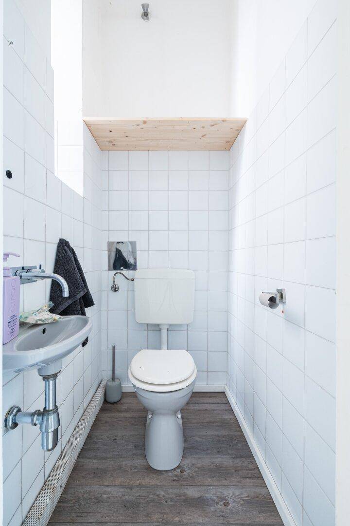 Kleines Gäste-WC mit Wand-WC, Waschbecken und Holzregal, hell gefliest.