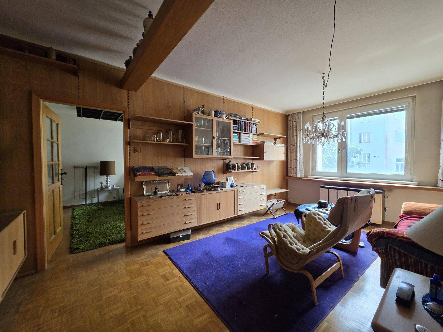 Geräumiges Wohnzimmer mit Holzwänden, Parkettboden und einem eleganten Kronleuchter, der den Raum erhellt.